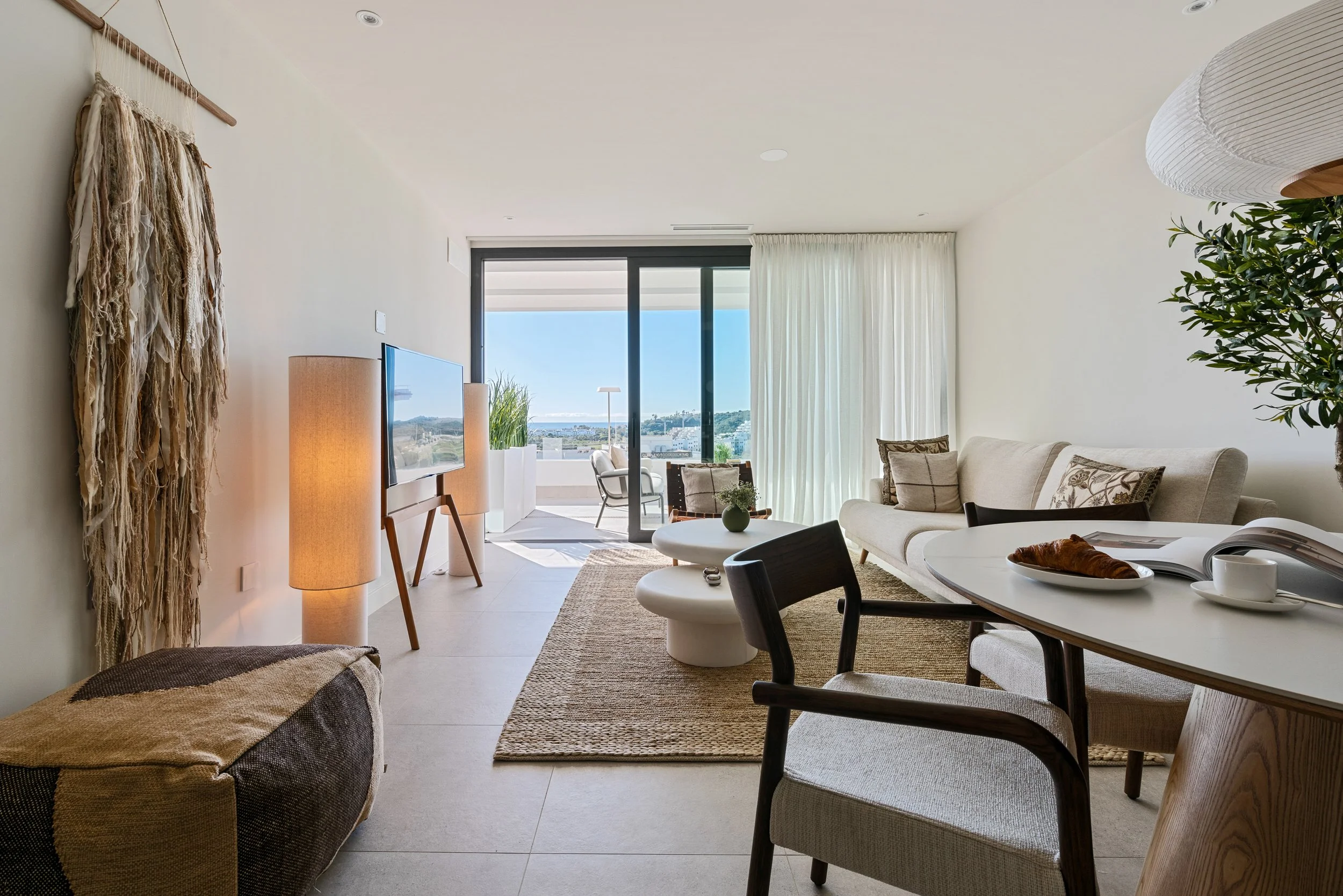 Living room with white walls, a large window with views of city and water, beige sofa, wooden chairs, a white round coffee table with a croissant and cups, a textured hanging wall art, a tall table lamp, and a sliding glass door leading to a balcony with outdoor seating.
