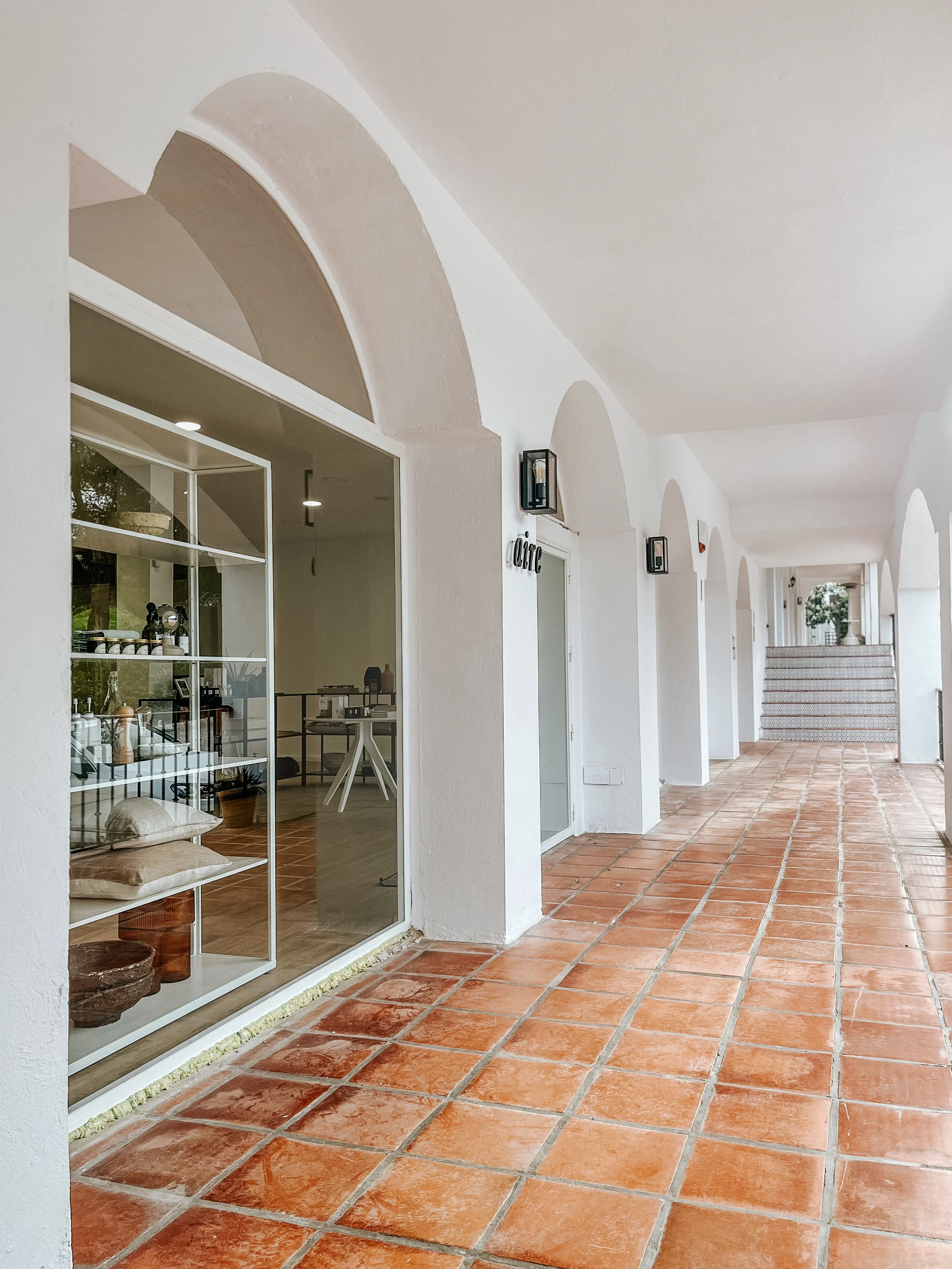 A covered outdoor walkway with white arches, terracotta tiles, and several glass doors leading to interior rooms. There are black wall-mounted lanterns along the hallway and potted plants at the far end.