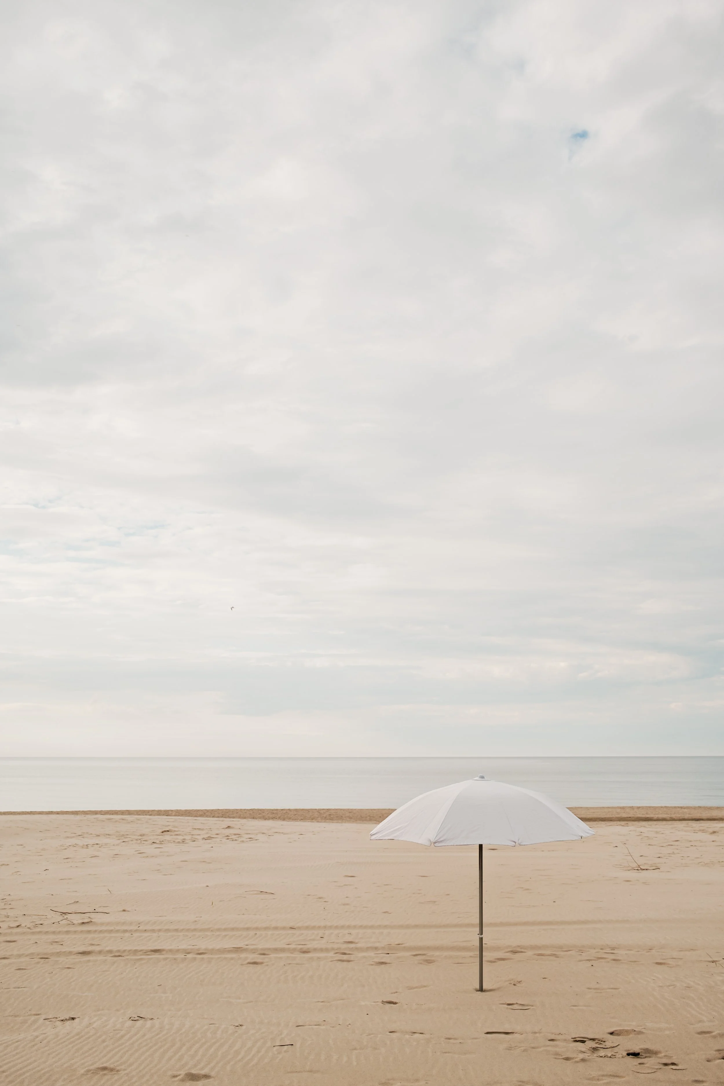 A white beach umbrella on a sandy beach with calm water and cloudy sky in the background.
