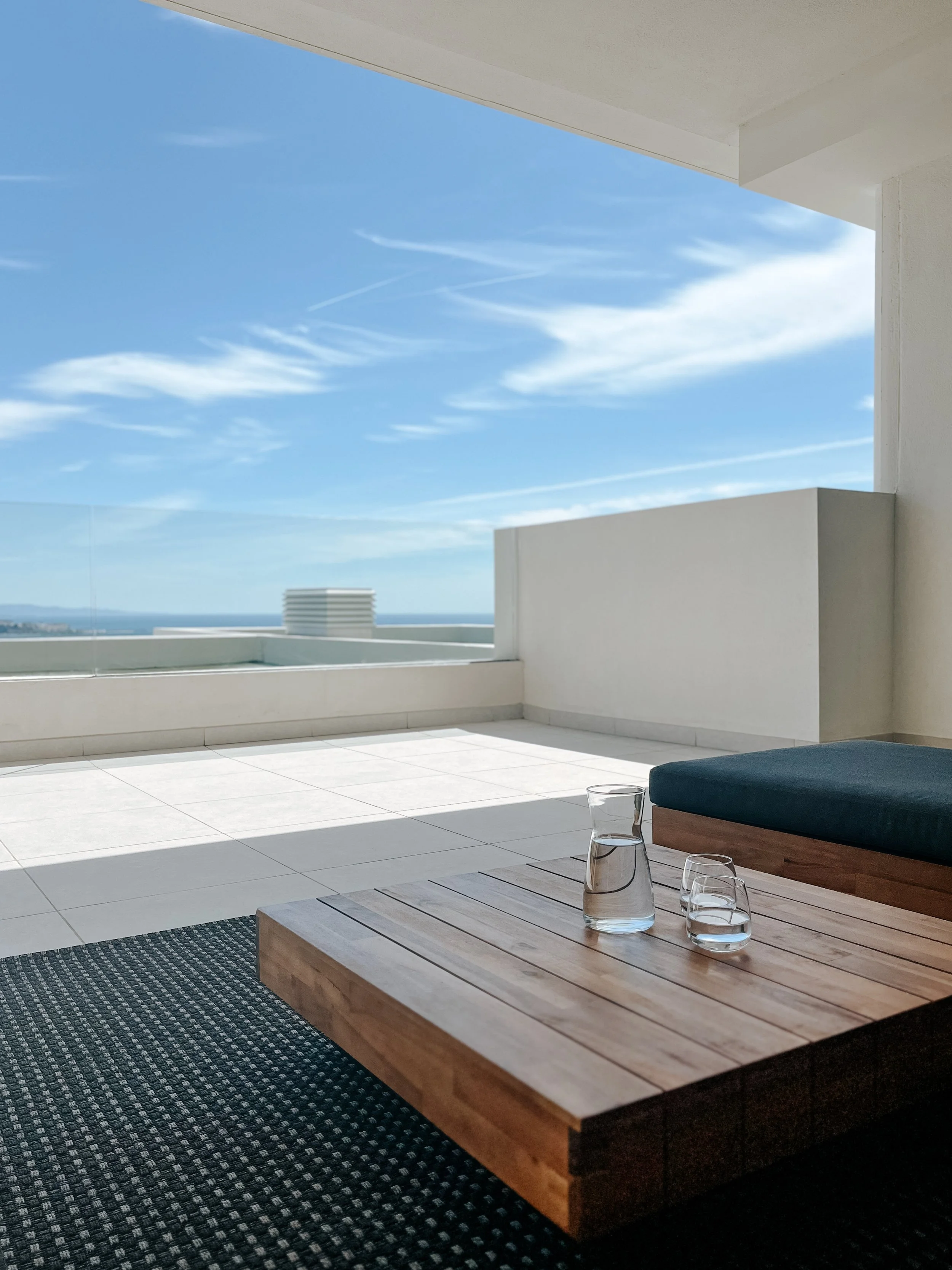 Modern balcony with white walls, glass railing, wooden coffee table, glass pitcher, and cups, with blue sky and clouds in the background.