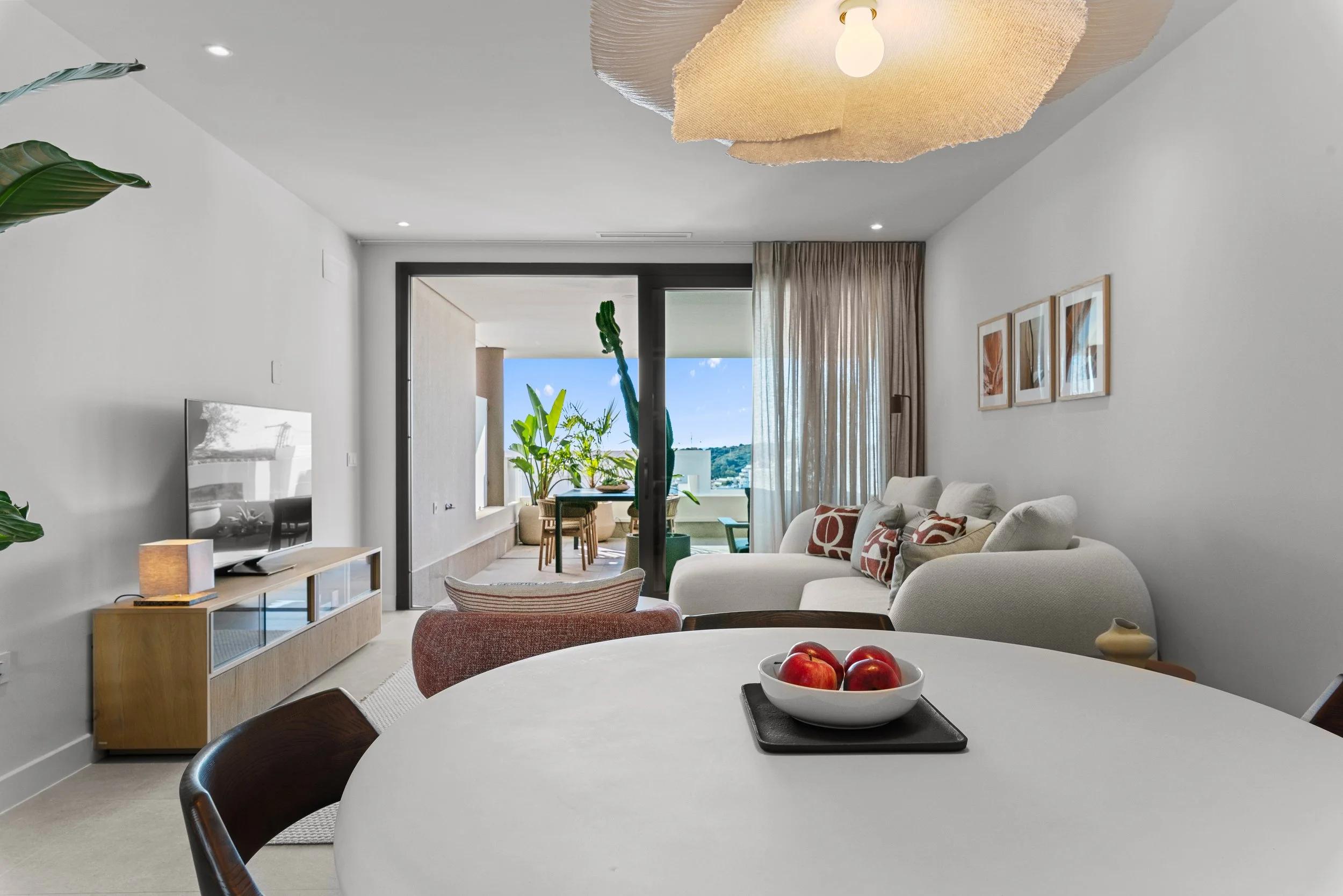 A modern living room with a white sectional sofa, patterned throw pillows, a dining area with plants outside, and a flat-screen TV on a wooden console.