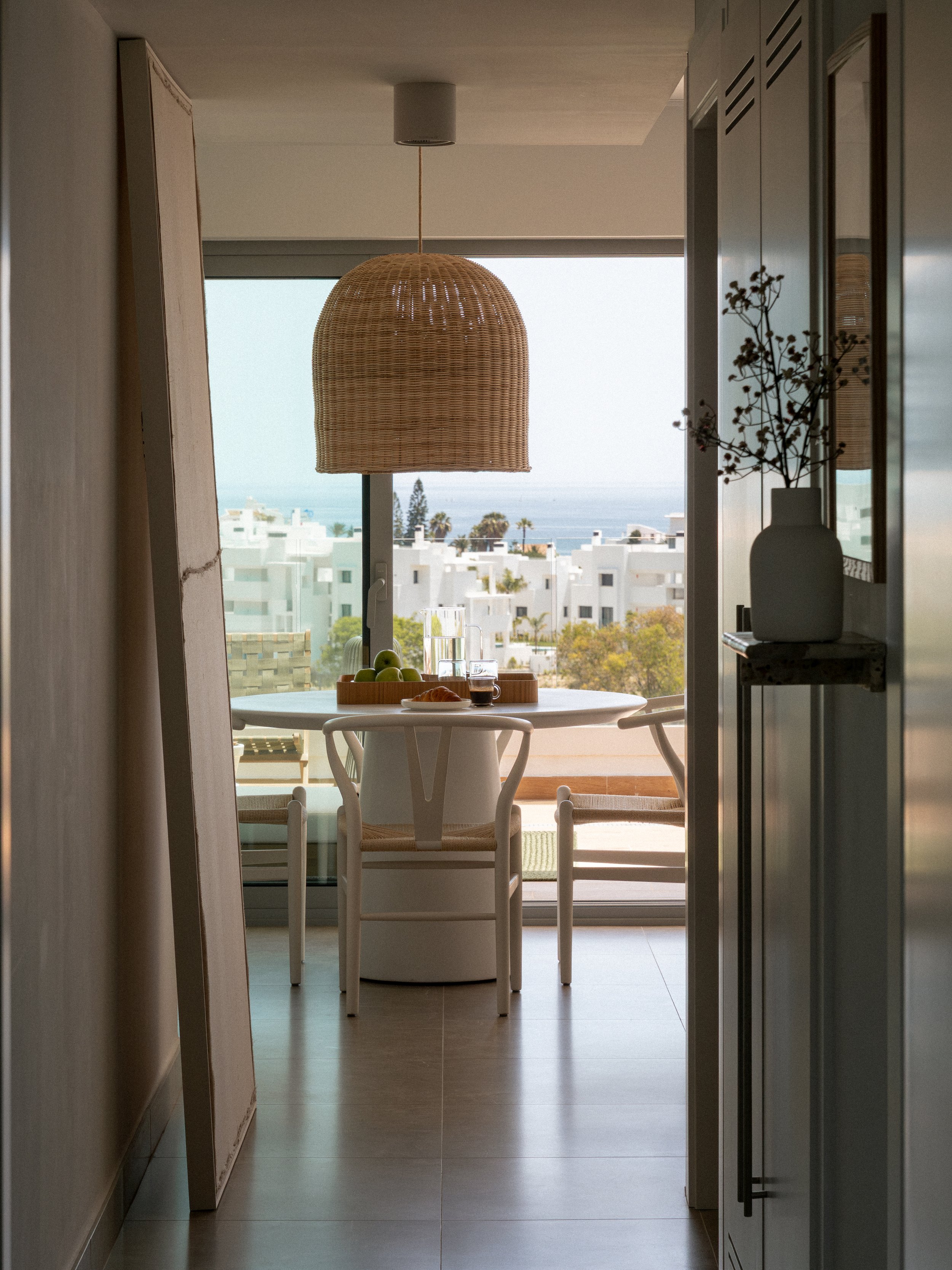 View from a hallway looking into a dining area with a round table and four chairs, decorated with a bowl of green apples, glasses, and a croissant, with a large window showing white buildings and palm trees outside, complemented by a woven pendant lamp hanging above.