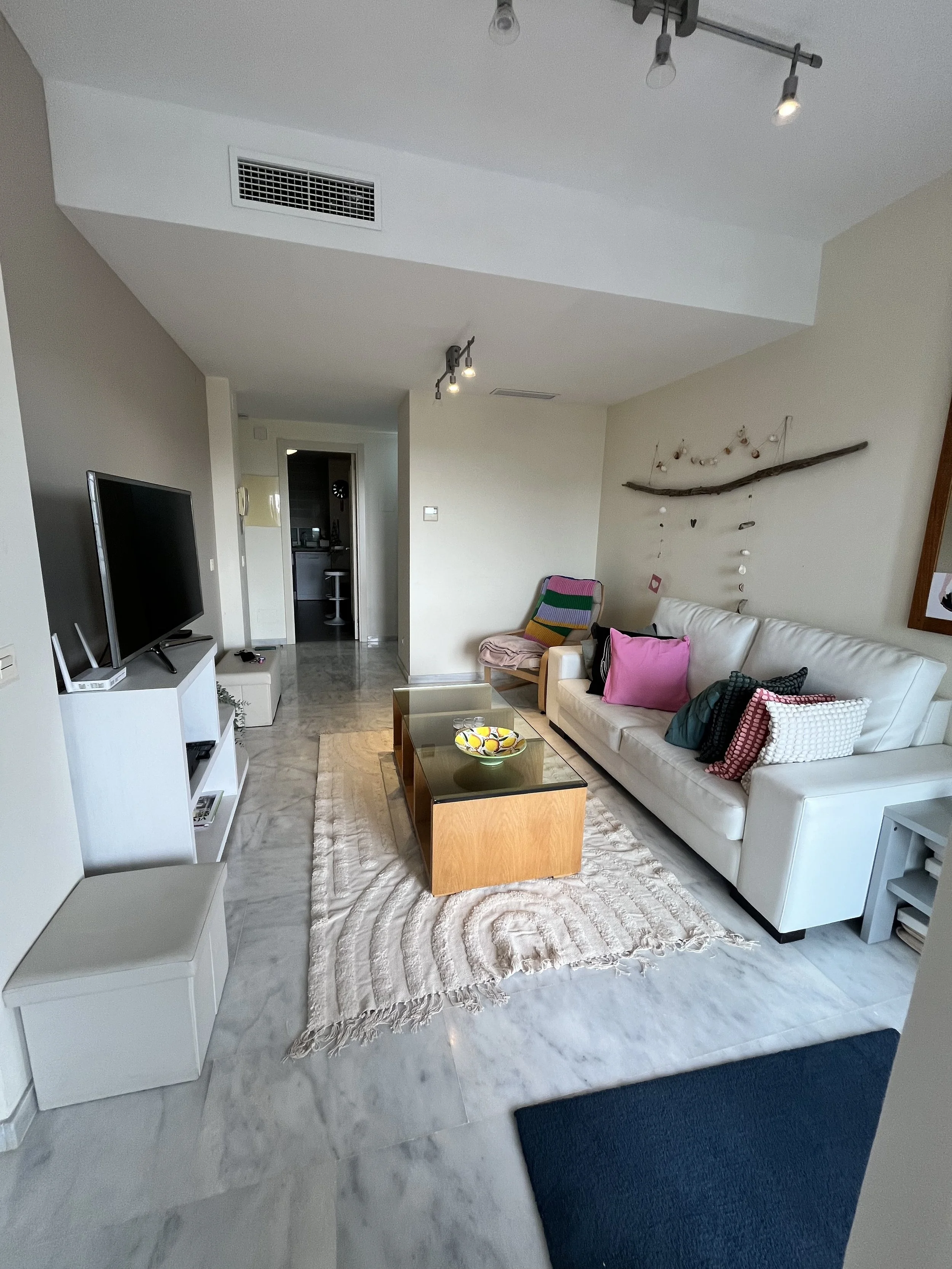 Living room with white sofa, colorful pillows, wooden coffee table with glass top, patterned rug, corner chair with multicolored knit blanket, wall decoration made of a branch and hanging shells, television on white stand, marble floor, ceiling track lighting, hallway leading to other rooms.