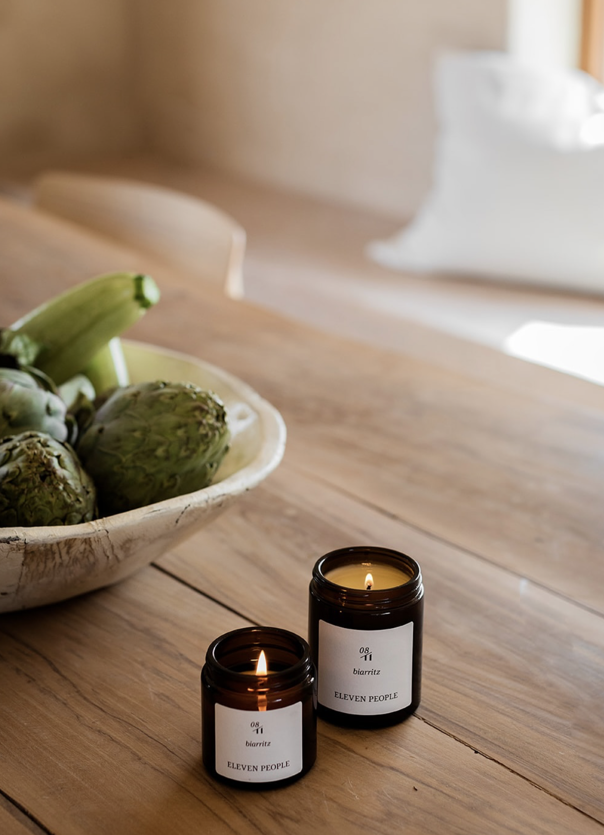A wooden table with two lit candles in brown glass jars labeled "ELEVEN PEOPLE," a bowl of artichokes, and a white cloth in the background.