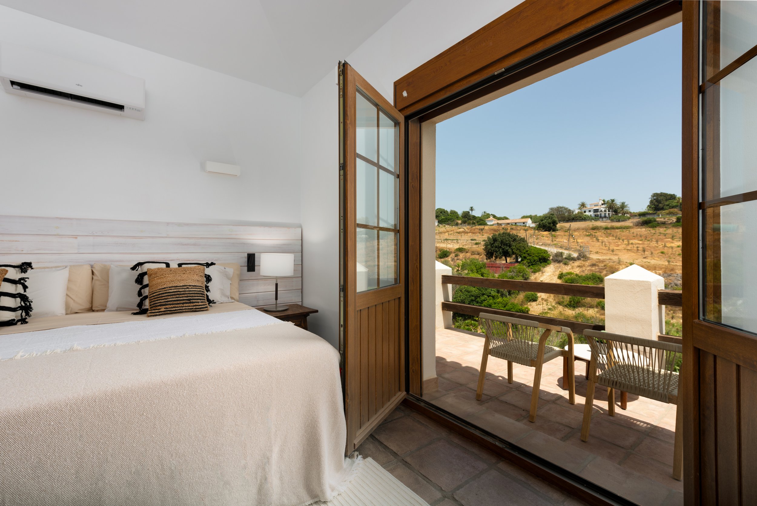 Bedroom with a large bed, beige bedding, and decorative pillows, open wooden balcony door showing a terrace with two chairs, overlooking a hilly landscape with trees and distant houses.