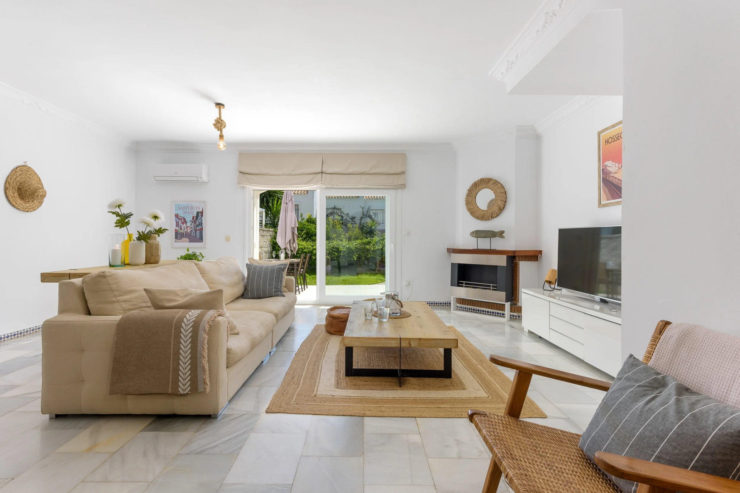 Living room with beige sofa, wooden coffee table, white entertainment unit with TV, decorative wall mirrors, artwork, and sliding glass door leading to a patio with outdoor furniture and greenery.
