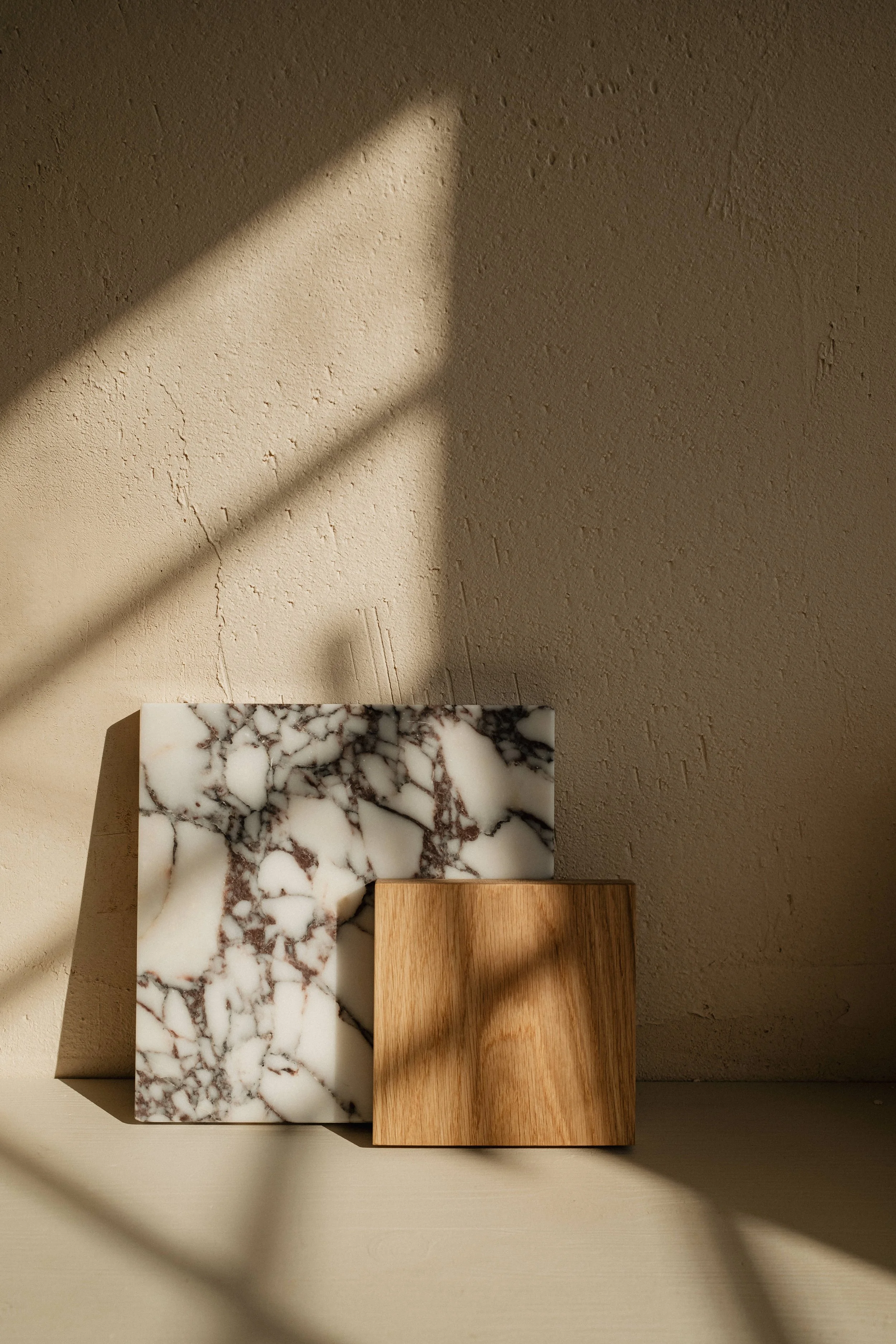 A photograph of two rectangular objects, one with a marble pattern and the other with a wood finish, placed on a surface against a textured beige wall. Shadows from a grid-like structure are cast across the objects and wall.