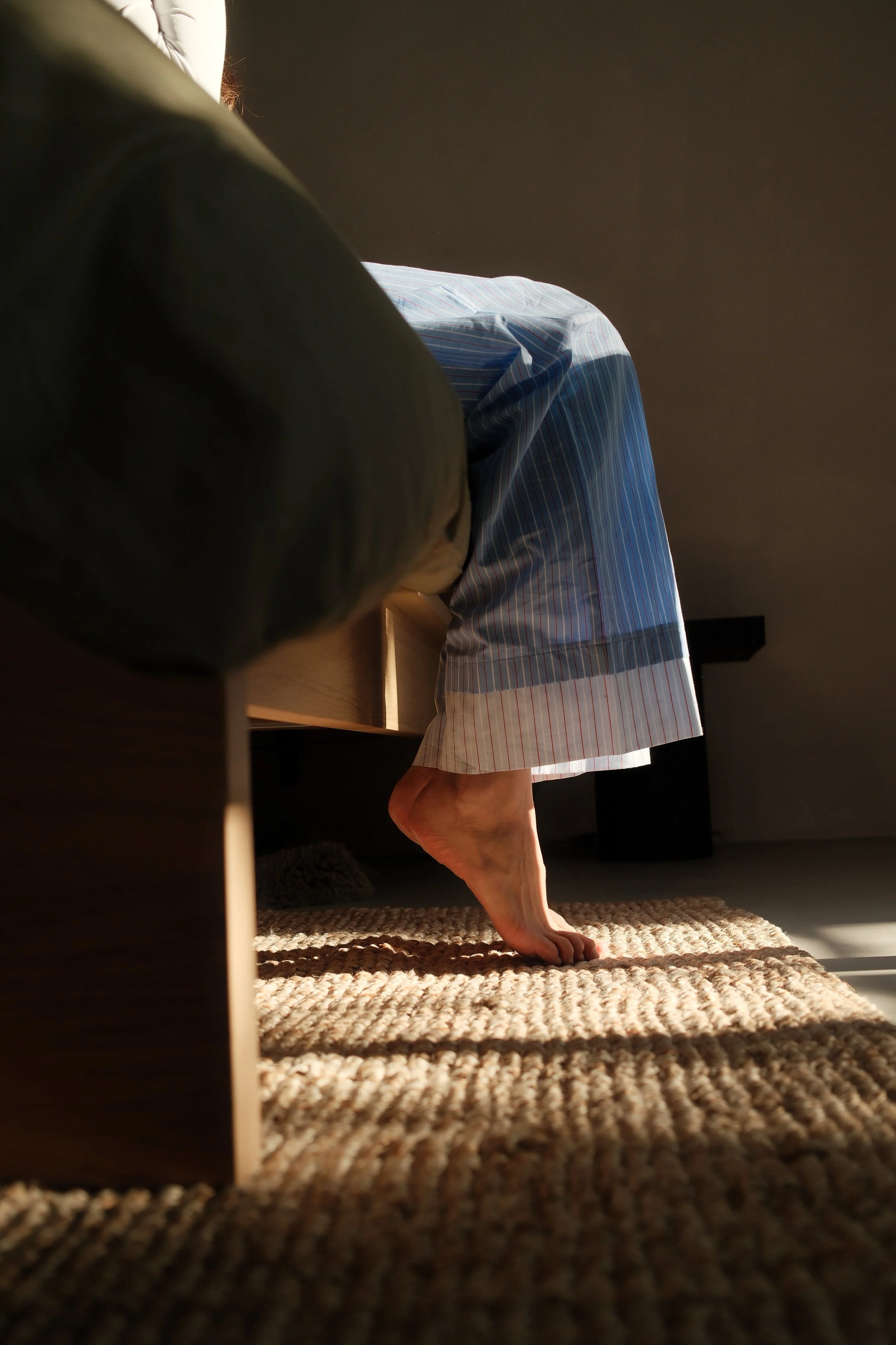 A person with one foot on a braided rug, wearing pajama pants, sitting on a wooden bed in a dimly lit room.