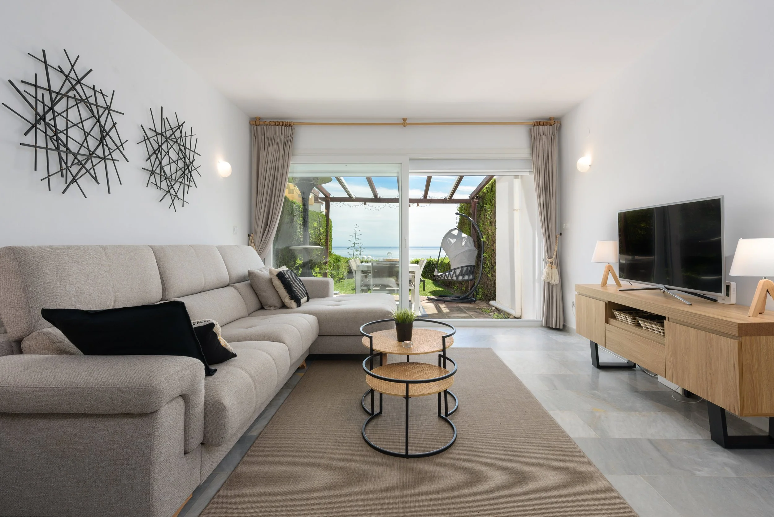 Living room with large beige sofa, coffee tables, and a TV on a wooden stand. Sliding glass door opens to outdoor patio with grass, table, and hanging chair with ocean view.