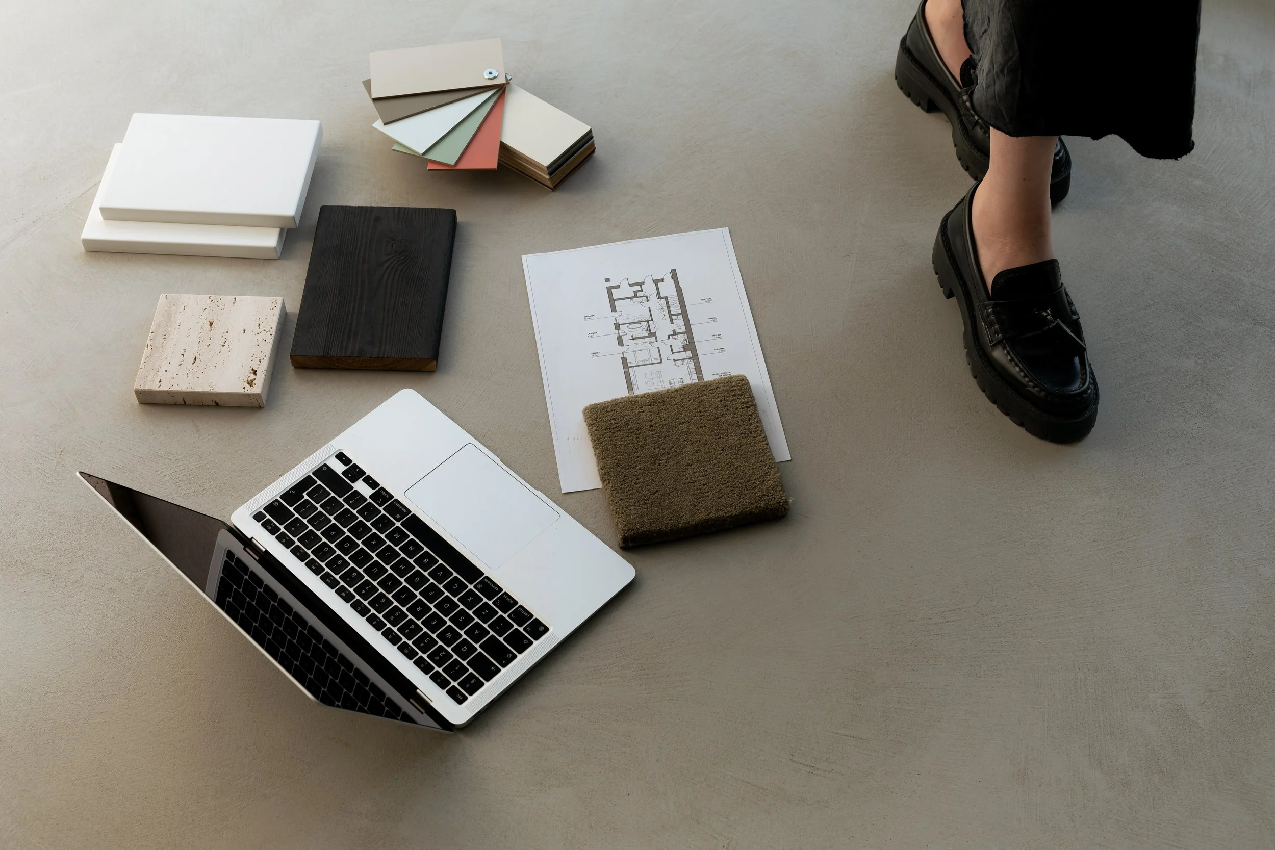Interior design or architecture workspace with samples of materials, a laptop, and a person wearing black shoes standing on a beige floor.