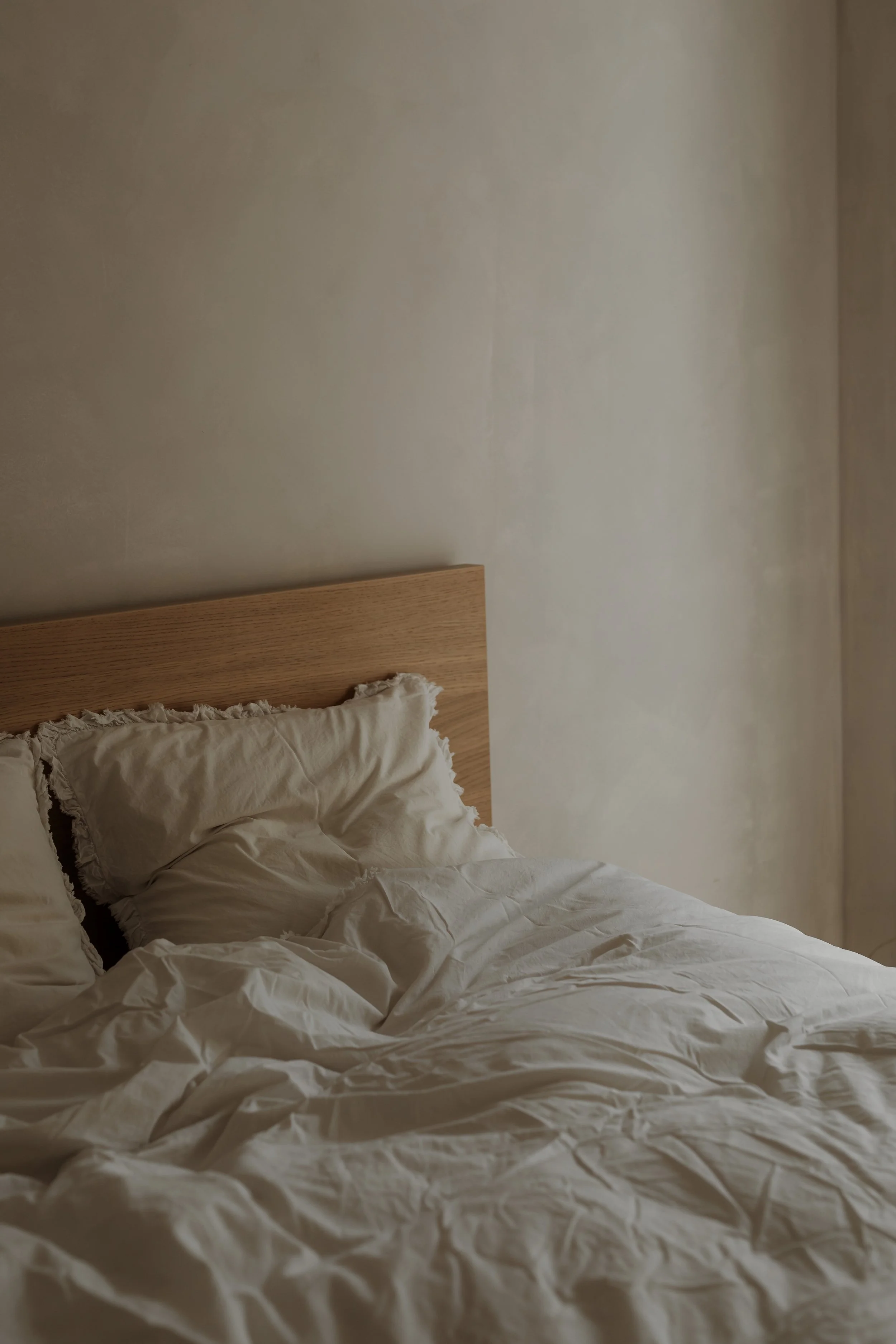 A neatly made bed with a wooden headboard, white crumpled bedding, and matching pillows in a softly lit bedroom.