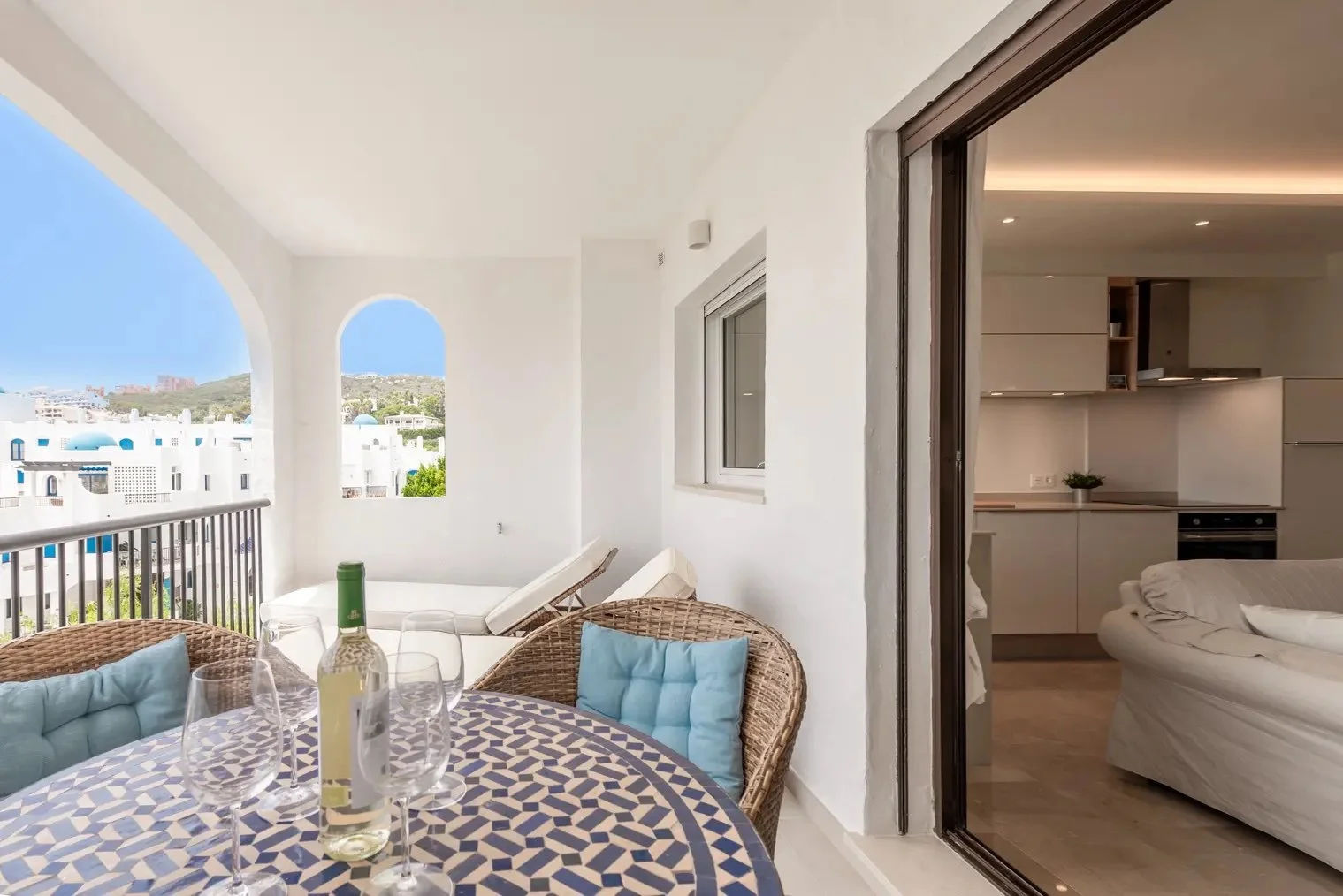 Balcony with a table set with wine glasses, a bottle, wicker chairs, and a view of white buildings with blue domes.