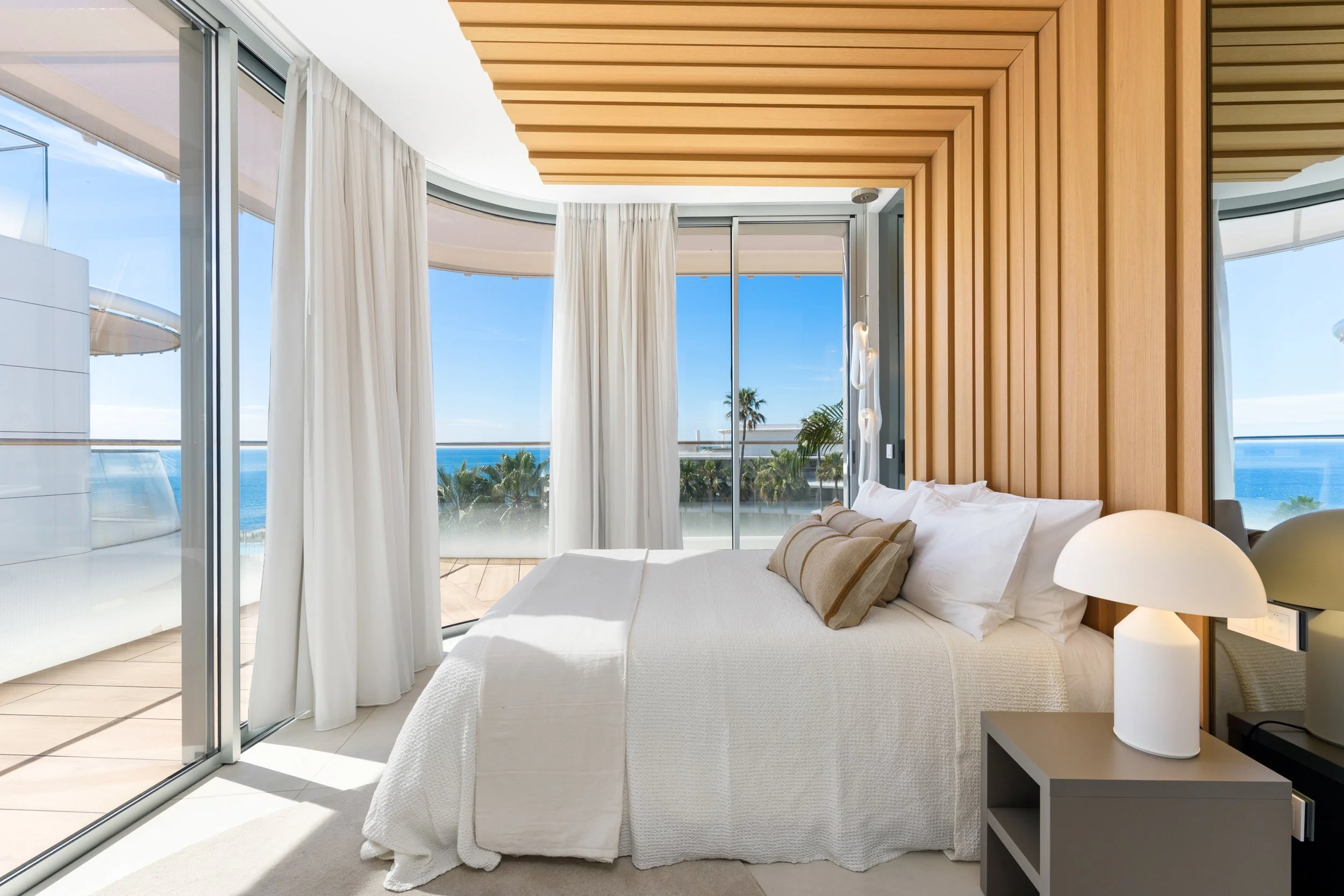 Bright hotel bedroom with large sliding glass doors overlooking a balcony, ocean and palm trees, decorated with a white bed, beige pillows, and a modern white table lamp.