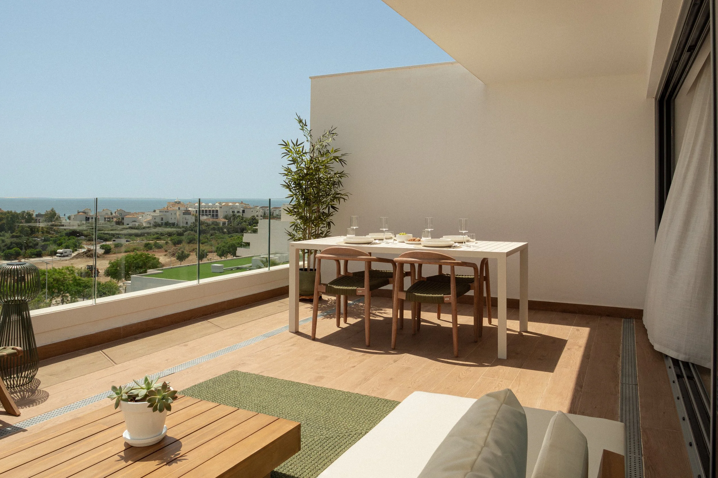 A balcony with a white dining table set for a meal, three wooden chairs, a small potted succulent on a wooden coffee table, and a view of houses, greenery, and the ocean under a clear sky.