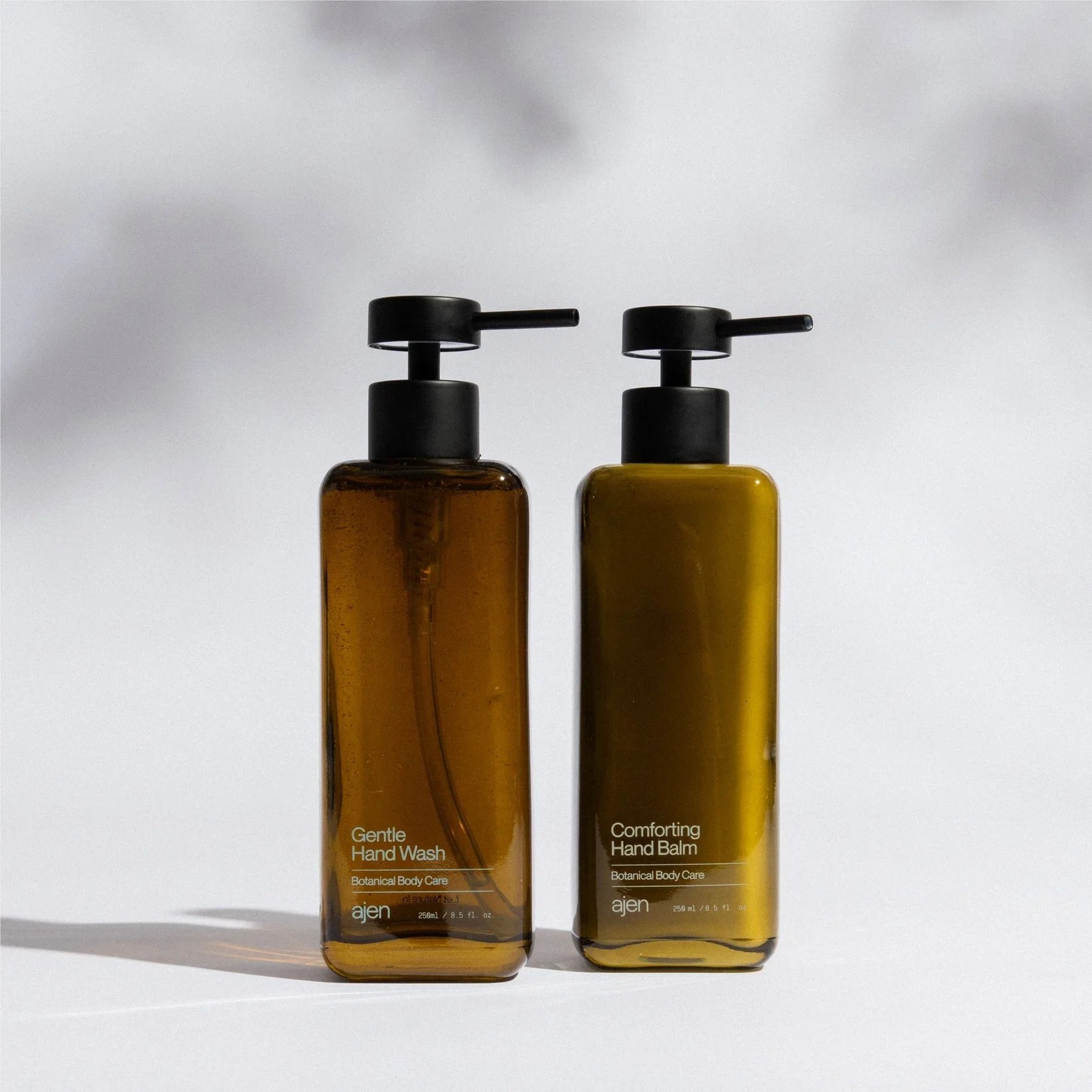 Two amber-colored bottles of botanical hand care products with black pump dispensers, labeled 'Gentle Hand Wash' and 'Comforting Hand Balm,' standing on a white surface with a light gray background.