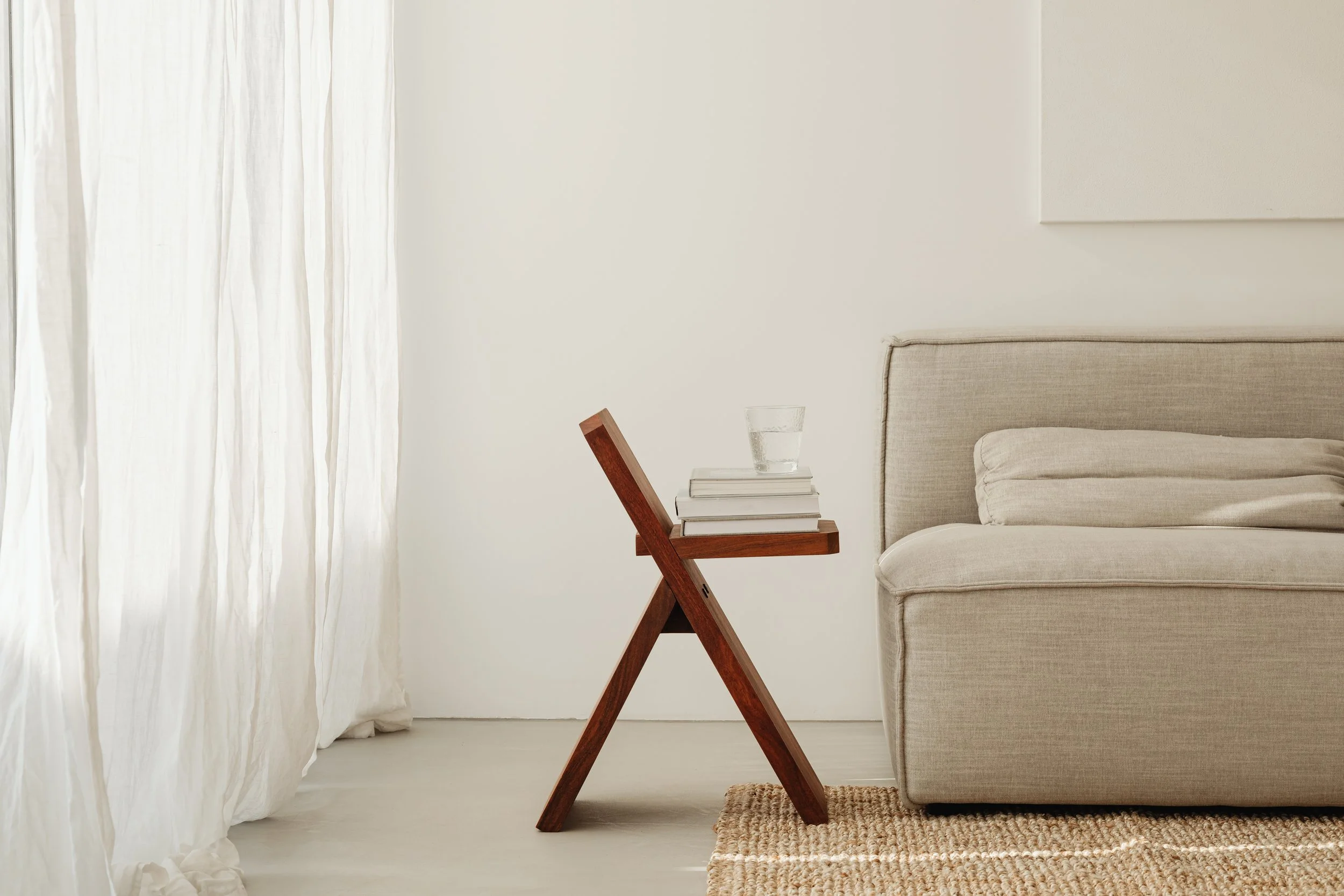 Living room corner with beige sofa, small wooden table holding books and a glass of water, and sheer white curtains.