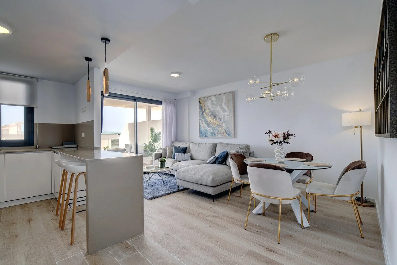 Open-concept living room and dining area with white walls, light hardwood floors, a beige sectional sofa, abstract wall art, a round dining table with four white and gold chairs, and a modern chandelier. The room has natural light from large glass sliding doors leading to a balcony with outdoor seating and plants.