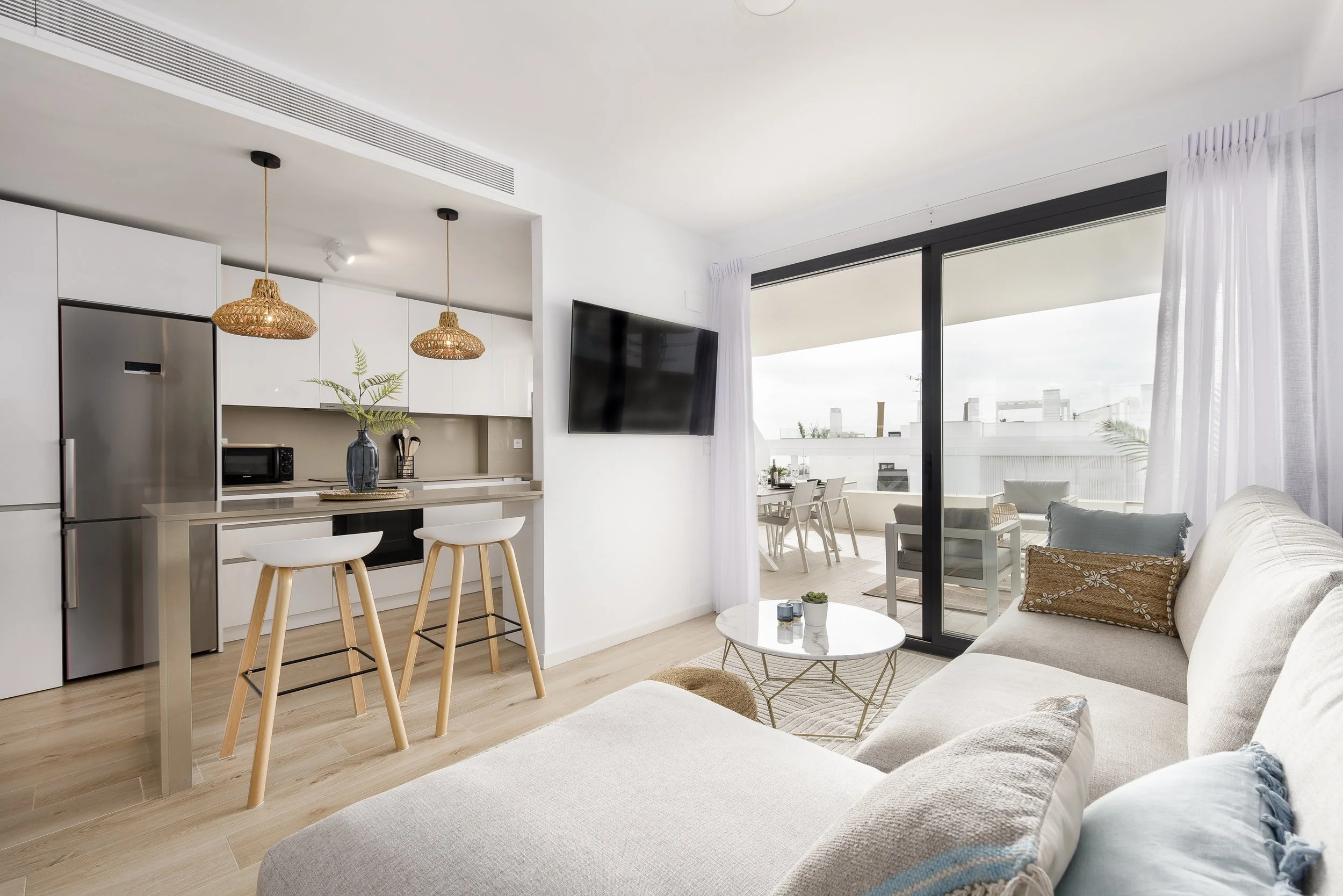 Modern living room with white sofa, glass sliding door leading to balcony, and kitchen area with white cabinets and bar stools