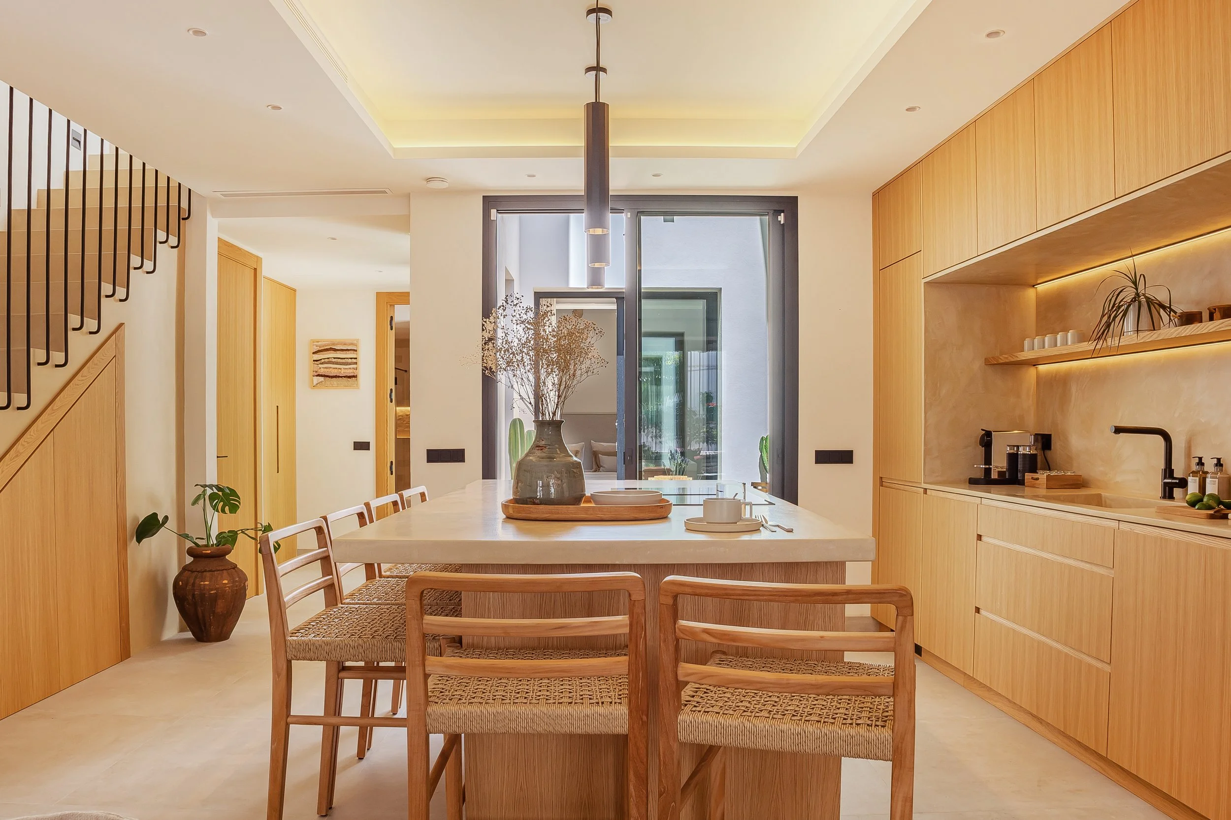 Modern kitchen and dining area with wooden cabinets, a central table with chairs, hanging pendant lights, and large windows with view of outdoor greenery.