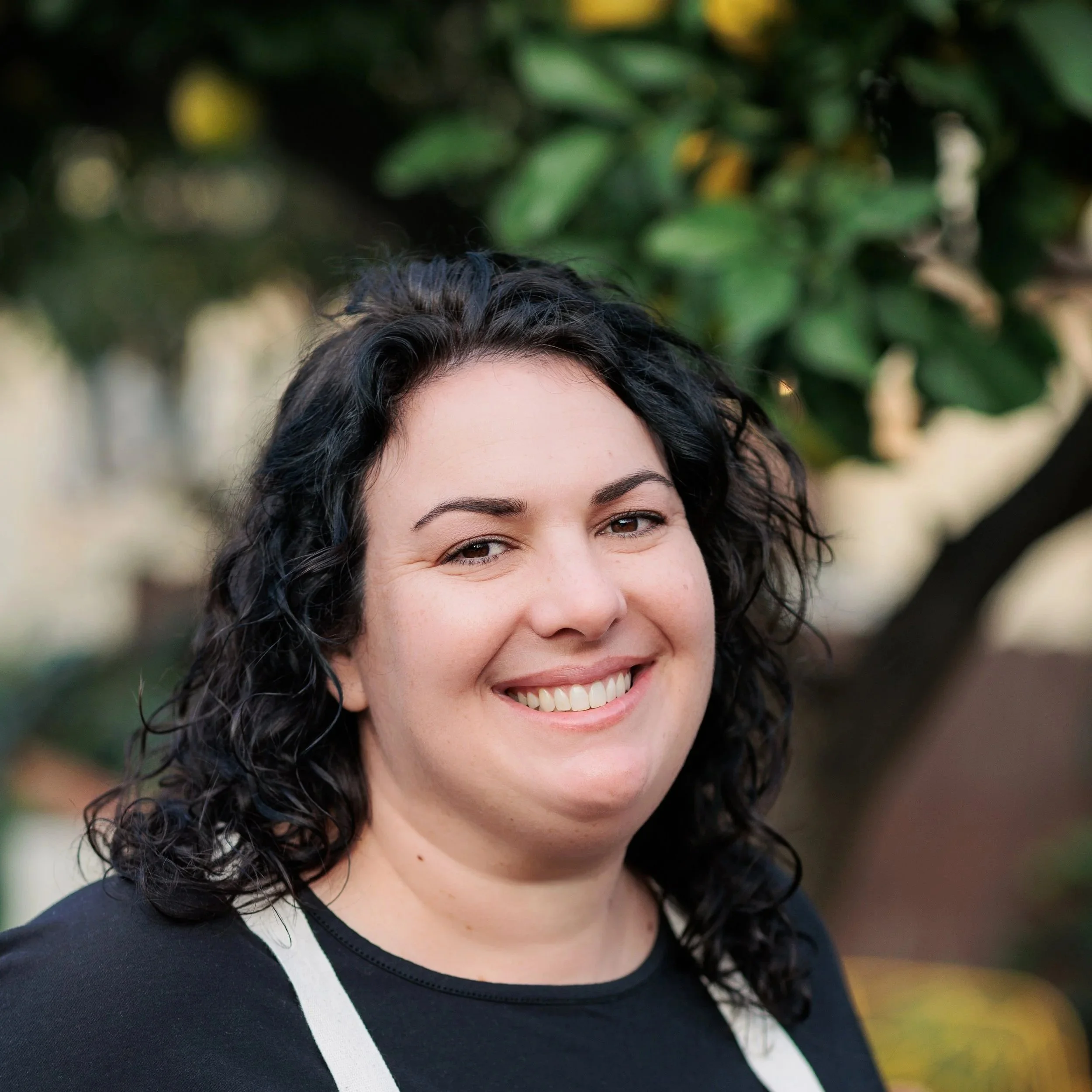 A woman with curly black hair smiling outdoors, with green foliage in the background.
