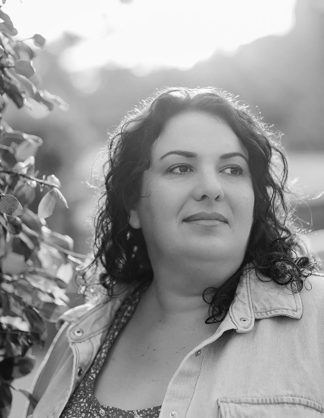 A black and white photo of a woman with curly hair, wearing a denim jacket over a patterned top, standing outdoors near trees and foliage with a soft, out-of-focus background.