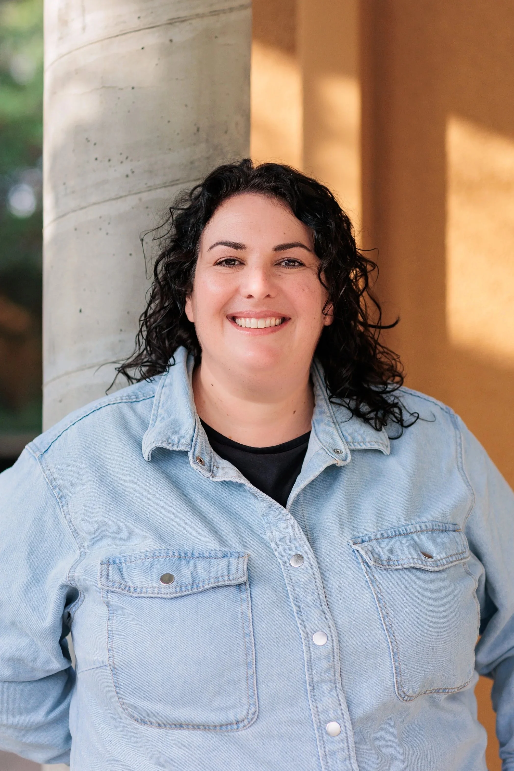 A woman with curly dark hair smiling, wearing a light denim jacket and black shirt, standing outside near a concrete pillar and a warm-colored wall.
