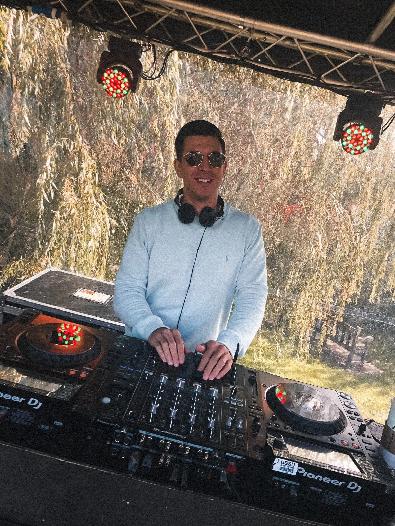 A DJ in sunglasses and headphones stands behind a mixing console outdoors, smiling.