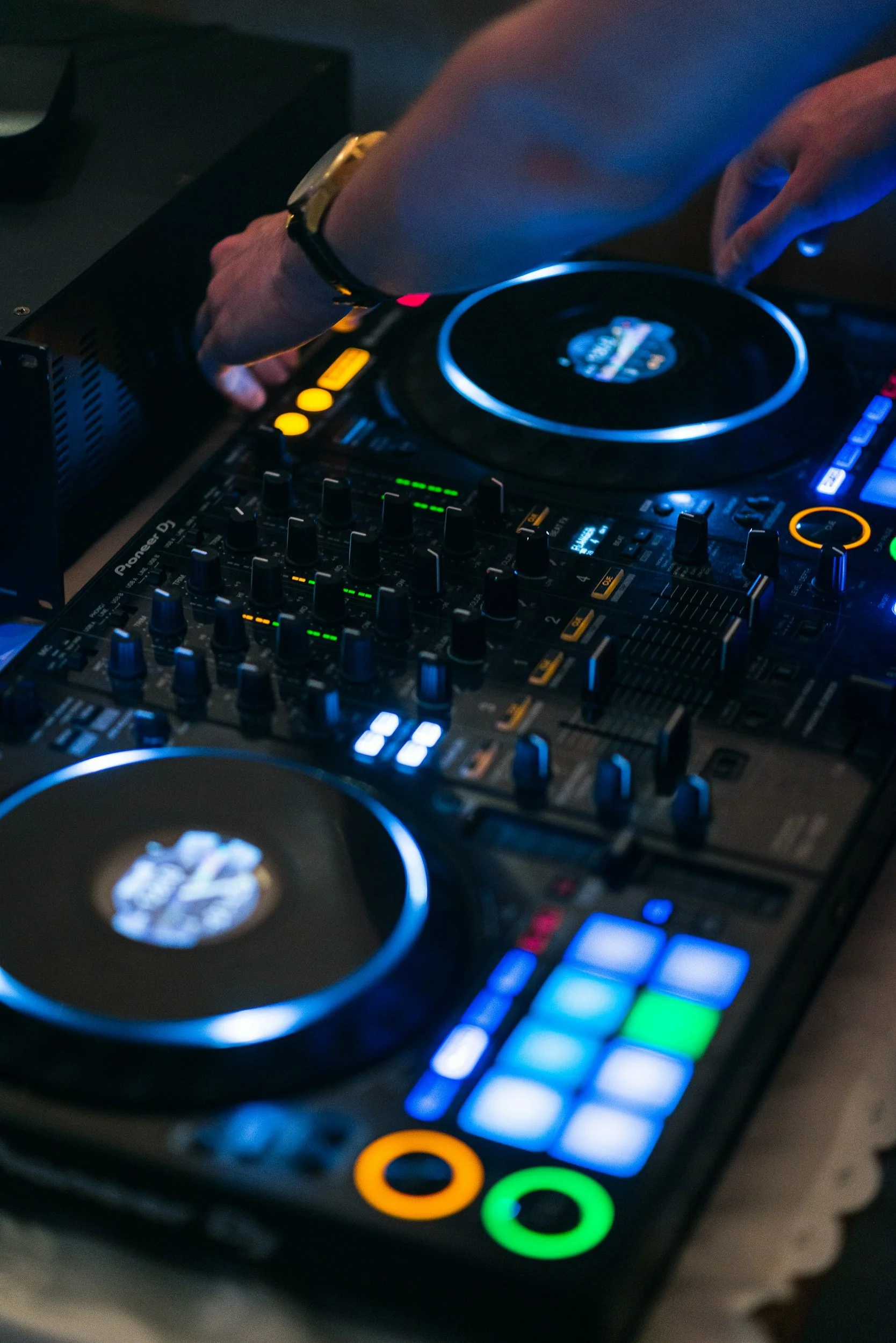 Close-up of a DJ's hands operating a professional DJ controller with illuminated buttons and knobs, in a dimly lit environment.