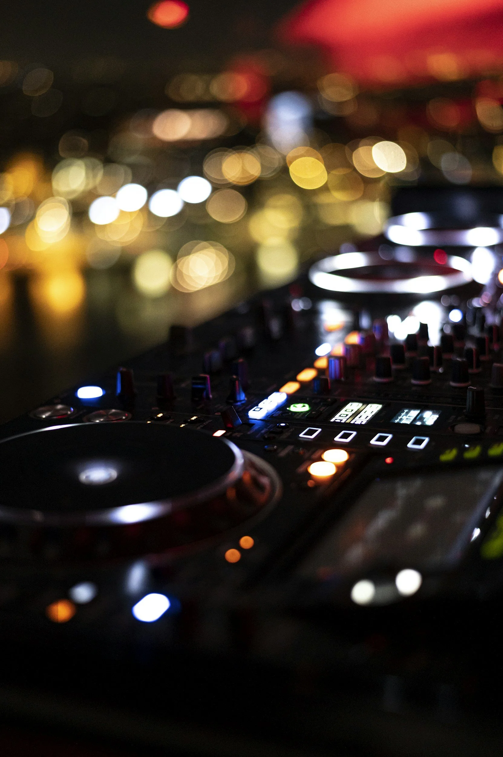 A DJ mixer and turntables with colorful LED lights against a backdrop of blurred city lights at night.