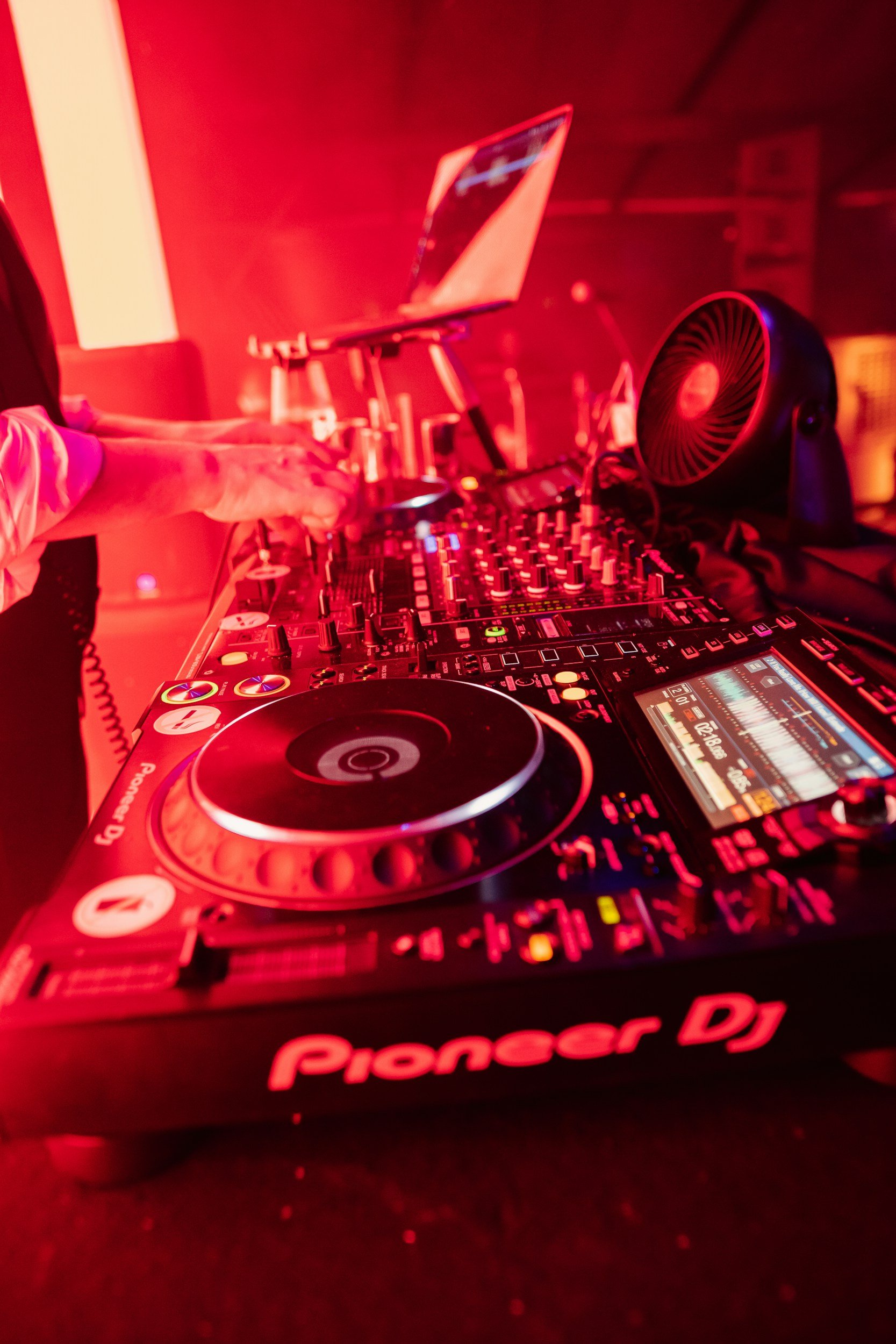 A DJ mixing music on a Pioneer DJ controller with a laptop in the background, in a club with red lighting.