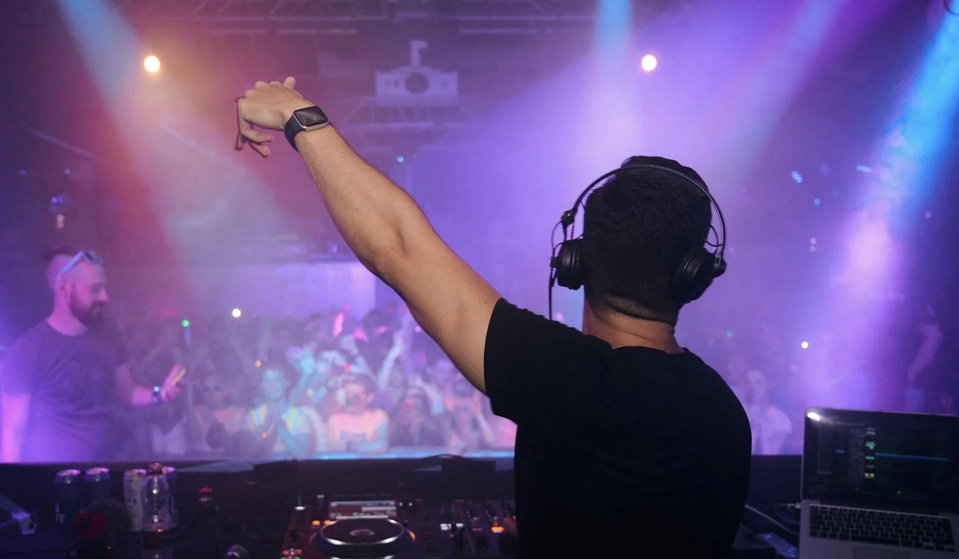 A DJ wearing headphones and a black shirt performing at a nightclub with a crowd dancing in the background, colorful lights, and a DJ controller and laptop on the table.