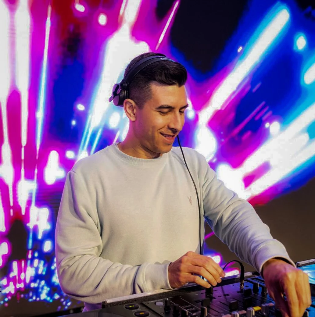 Male DJ wearing headphones, smiling, operating DJ equipment with colorful light display in background.