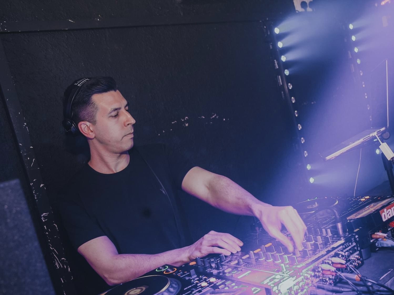 A male DJ wearing headphones operates DJ equipment during a performance, with blue and purple stage lights illuminating the scene.
