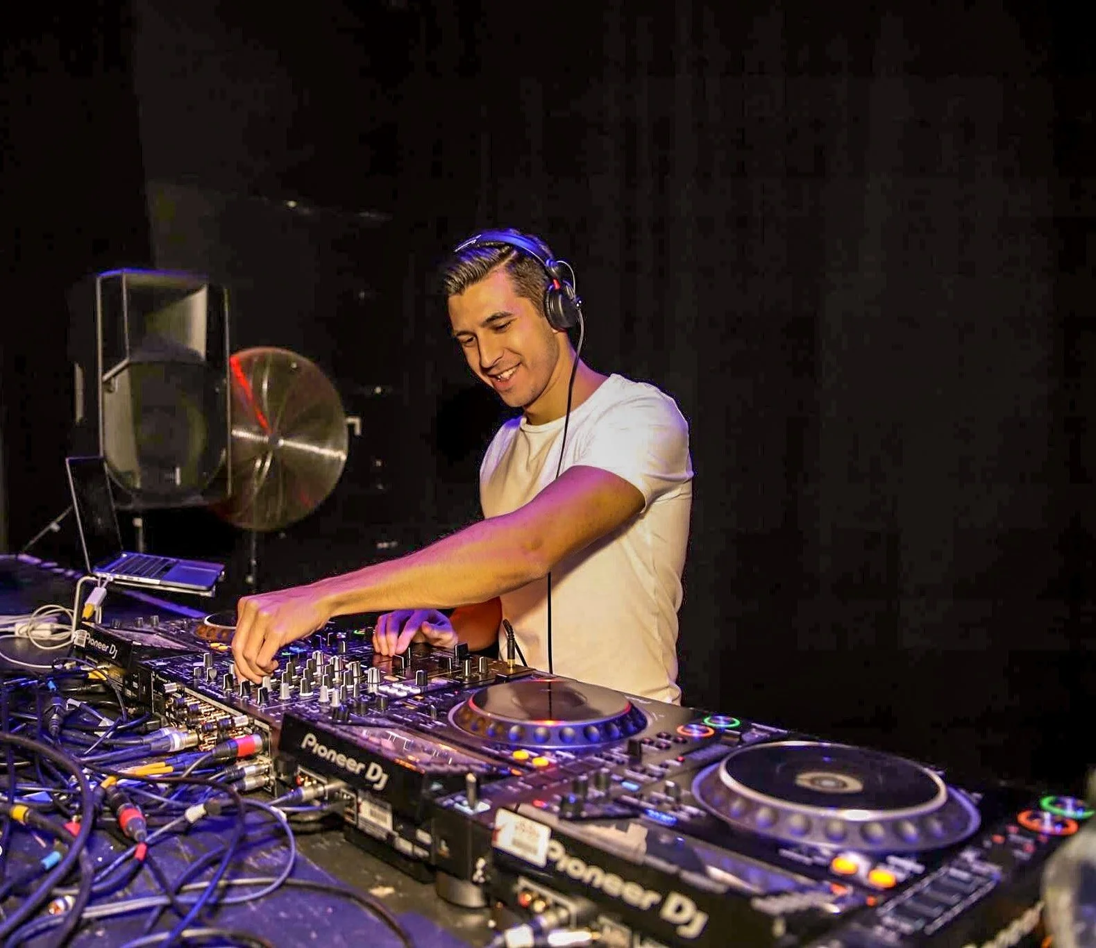 A DJ smiling and wearing headphones, standing behind a DJ mixer and turntables, in a dark club setting.