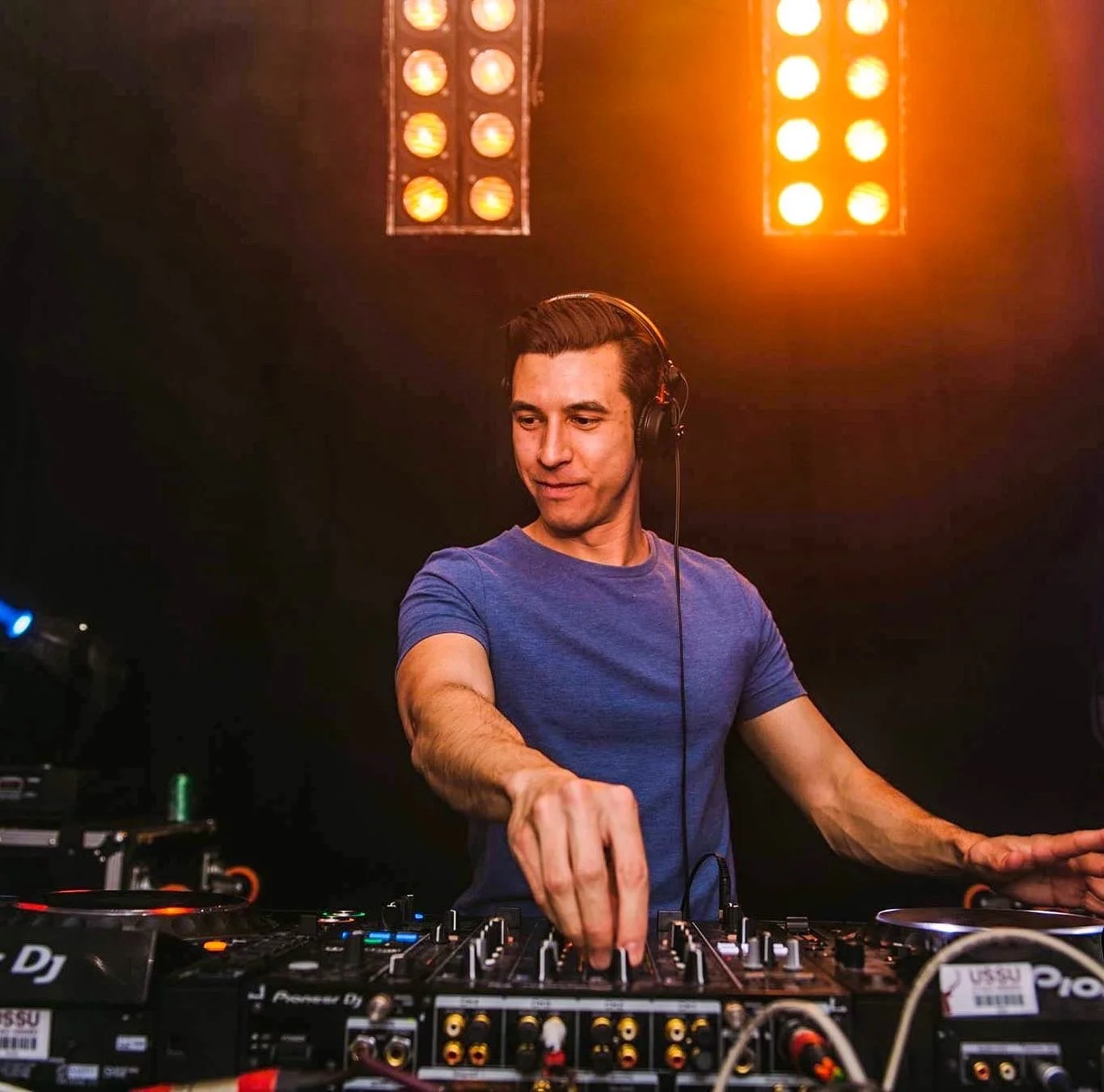 A male DJ wearing a blue t-shirt and headphones is mixing music on a DJ console in a dark setting with orange stage lights in the background.