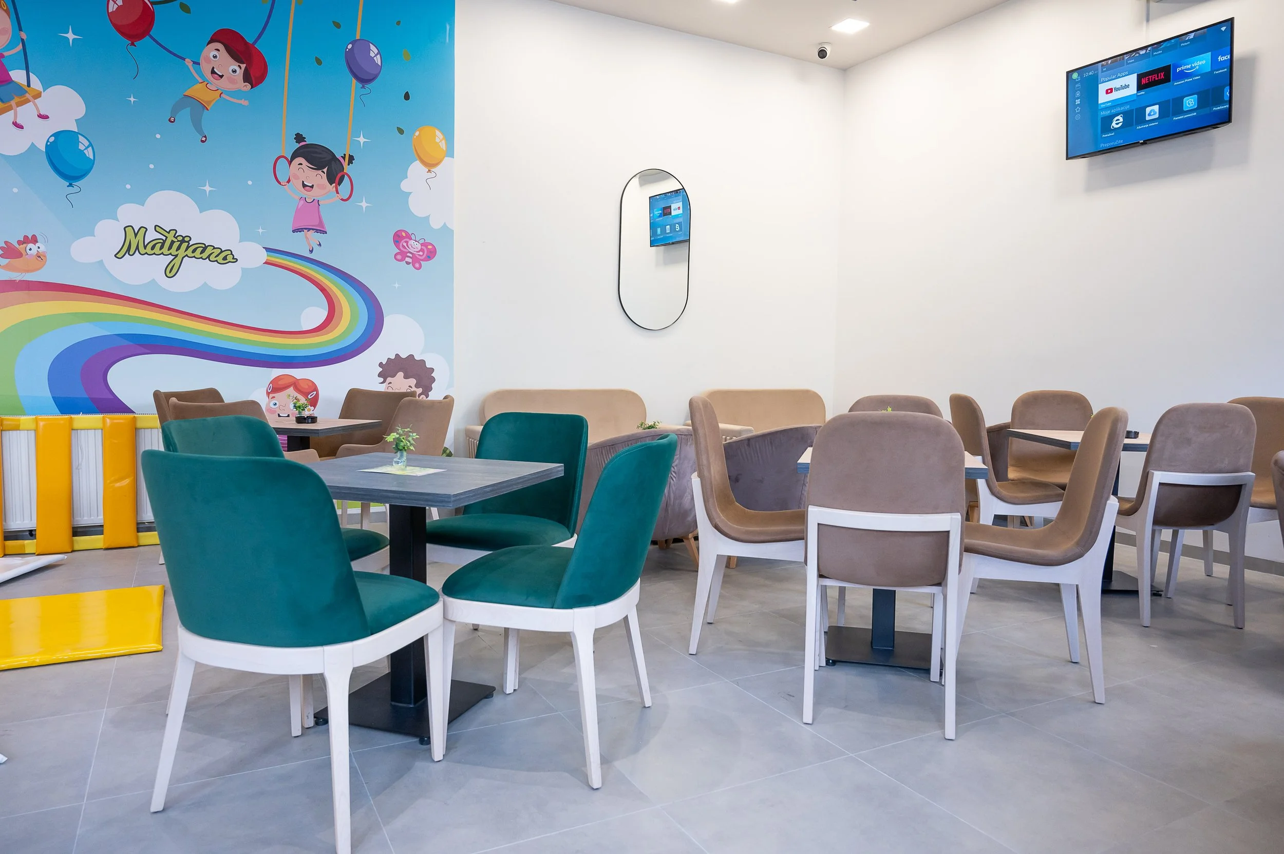 A brightly lit, modern children's play area or family lounge with colorful cartoon characters on a wall mural, small round tables, and upholstered chairs in teal, beige, and gray tones. A television is mounted on the wall, and a mirror is on the white wall.