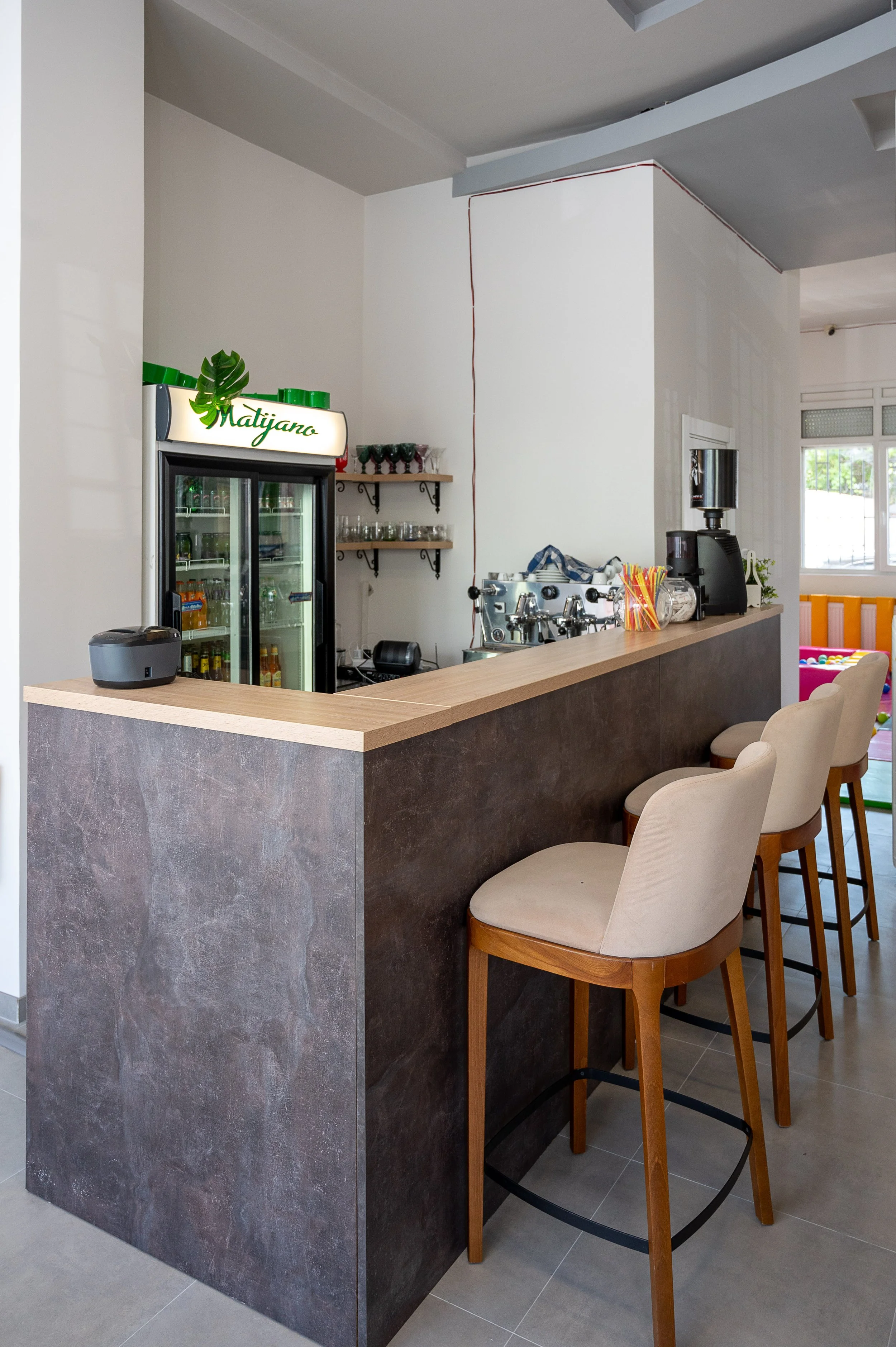 Interior view of a modern bar area with beige bar stools, a black bar counter with a wooden top, a drink refrigerator, and coffee machines. There's a small shelf with glasses and condiments, and a bright window in the background.