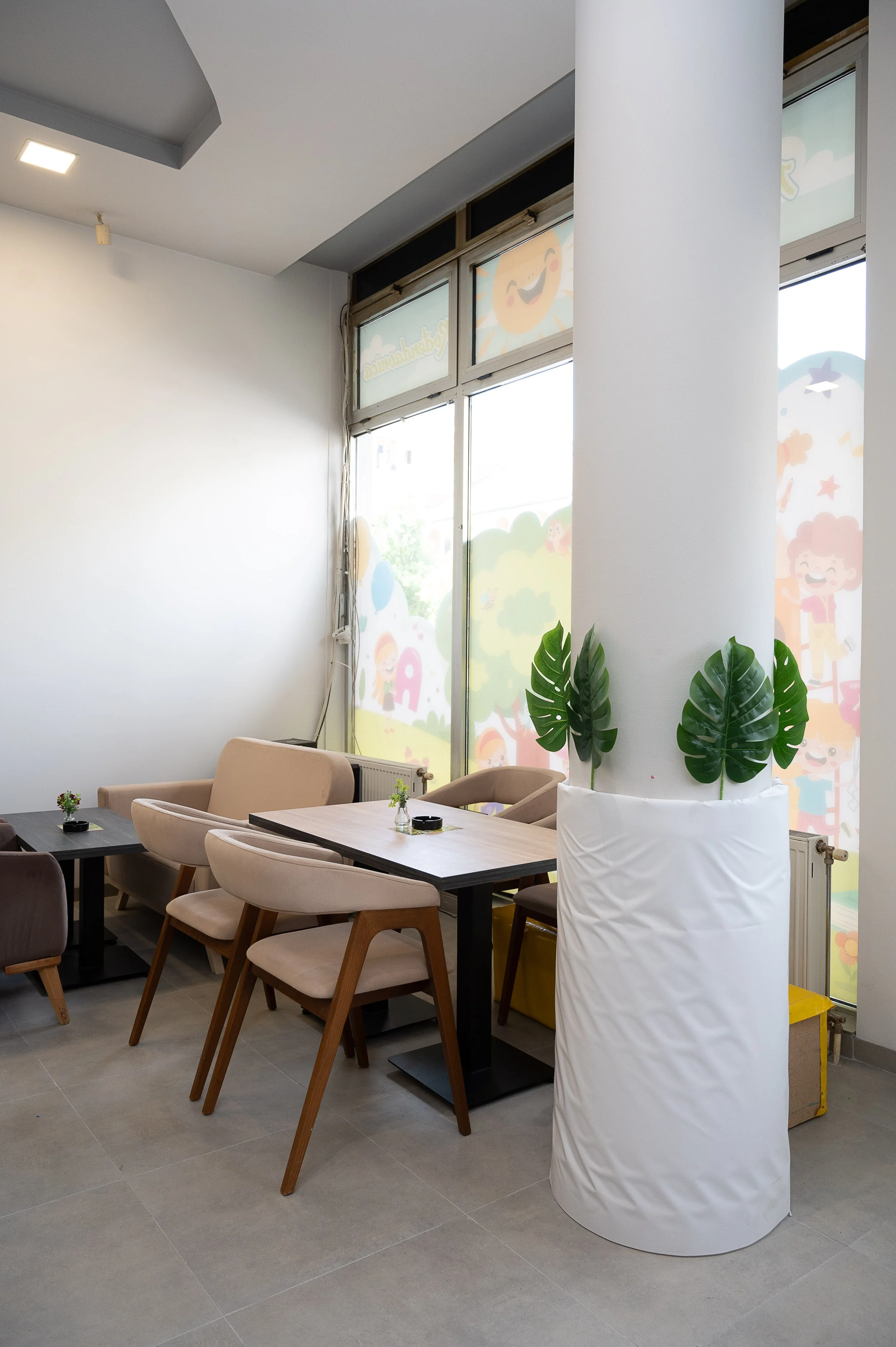 Interior of a cozy cafe with beige and brown chairs, wooden tables, small vases with flowers, large window with colorful cartoon cloud and character decorations, and a white potted plant.
