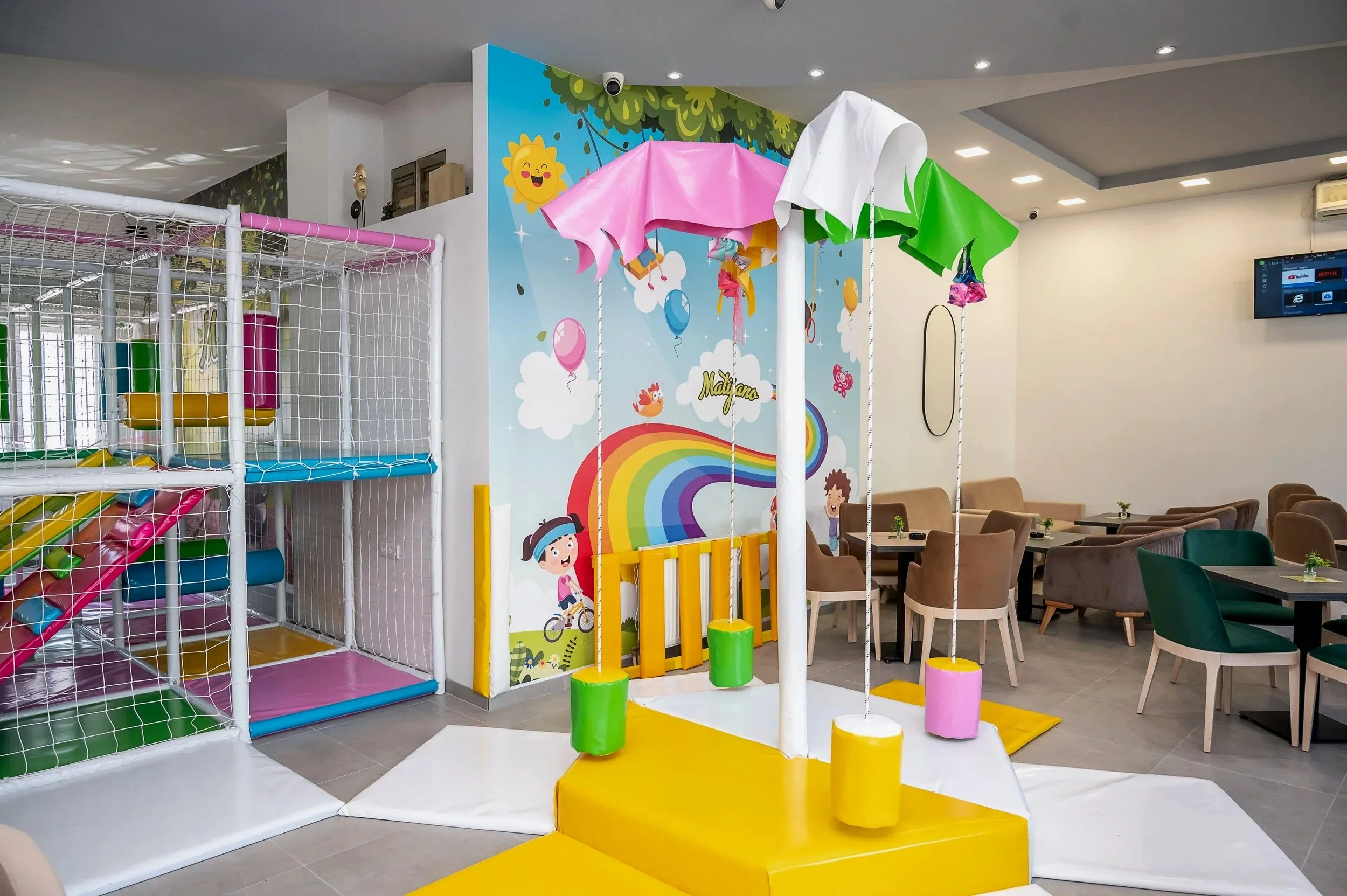 Indoor children's play area with colorful swings, a ball pit, climbing structures, and a cartoon mural with a rainbow, clouds, and children on the wall