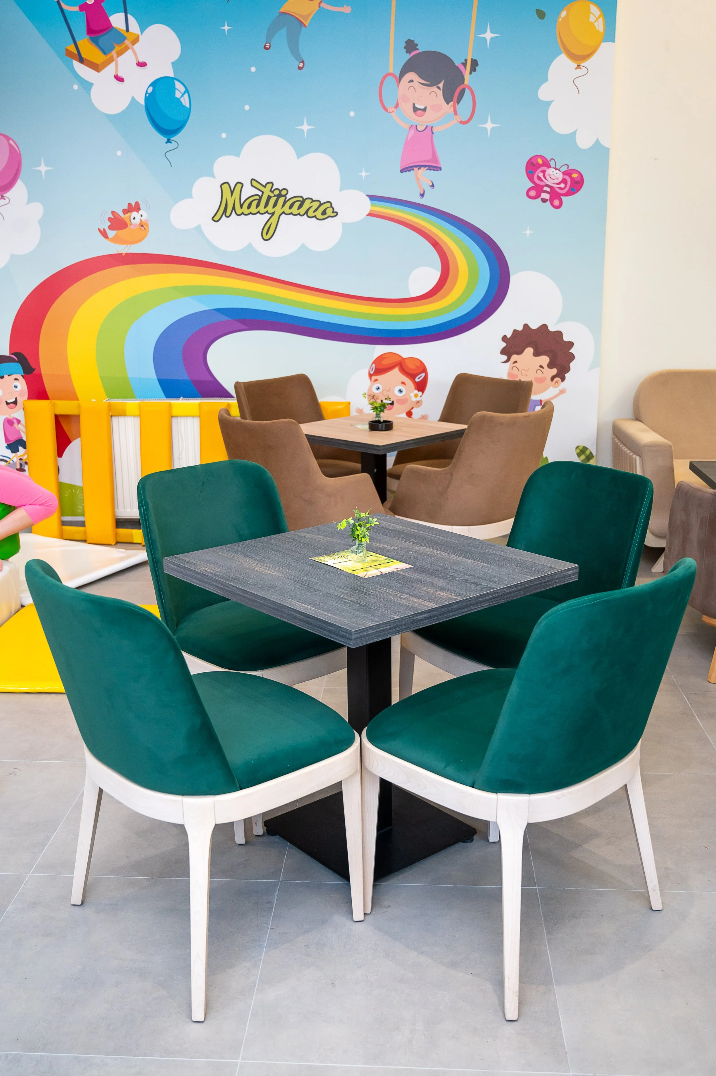 Interior of a colorful children's play area with tables and chairs, featuring a vibrant wall mural of a rainbow, cartoon children, animals, and clouds.