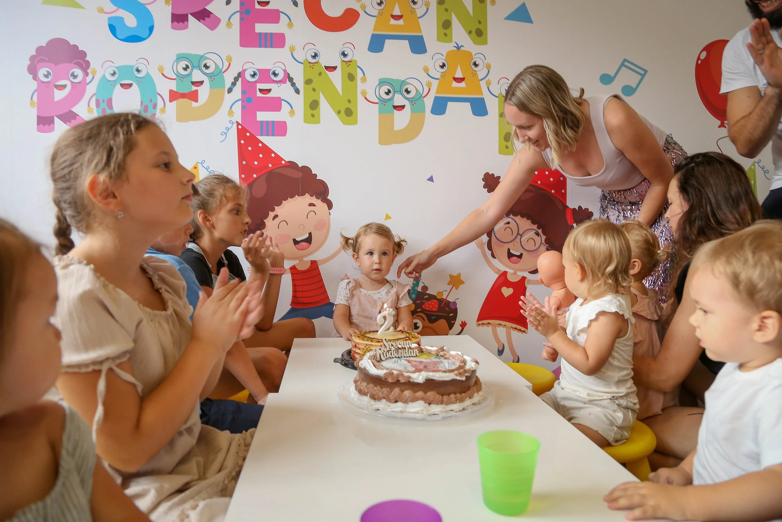 Children gathered around a birthday cake with a sister or friend in front of a decorated party backdrop with colorful letters and cartoon characters, celebrating a second birthday.