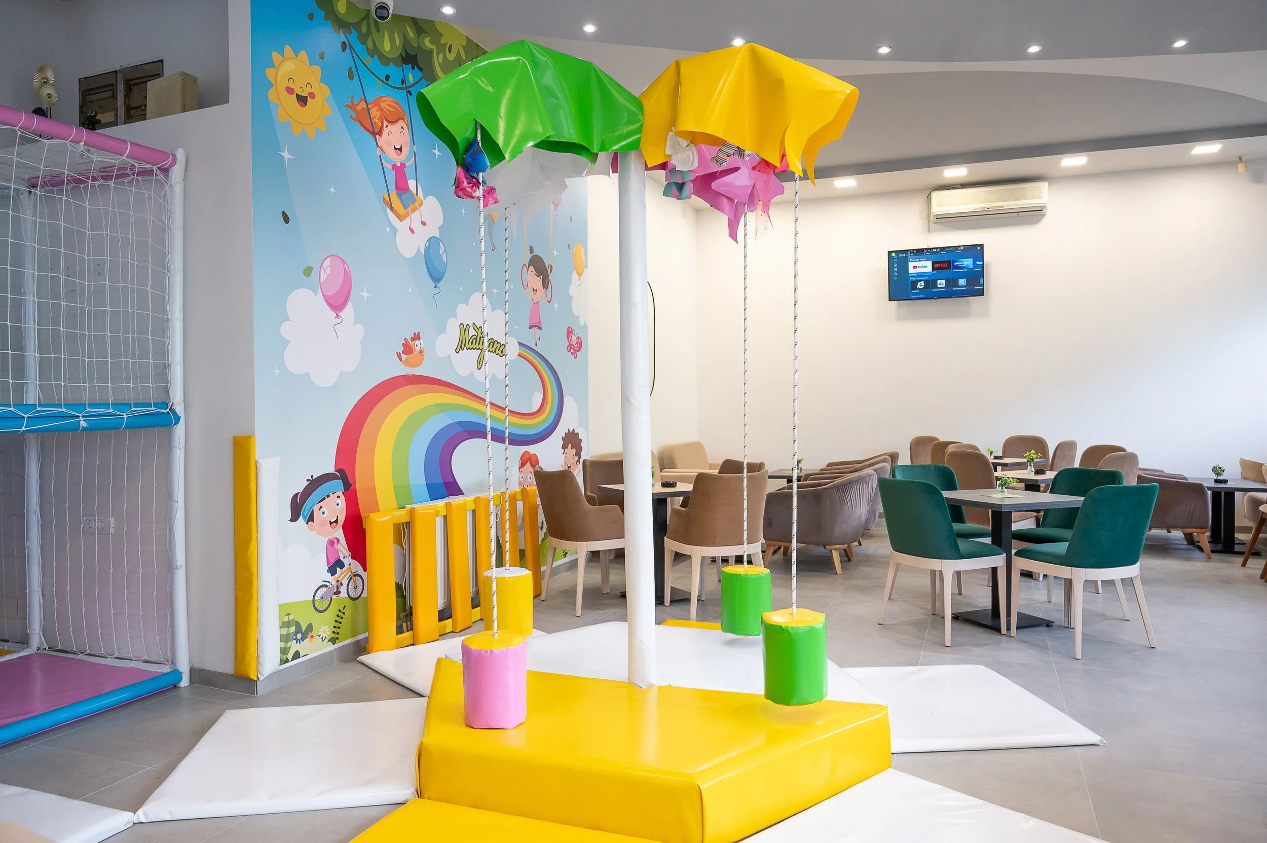 Colorful indoor play area featuring a small carousel with four matched seats, each with a colorful canopy in pink, green, yellow, and darker pink. The backdrop has a cartoon mural of children, a rainbow, and cheerful weather elements. To the left, th