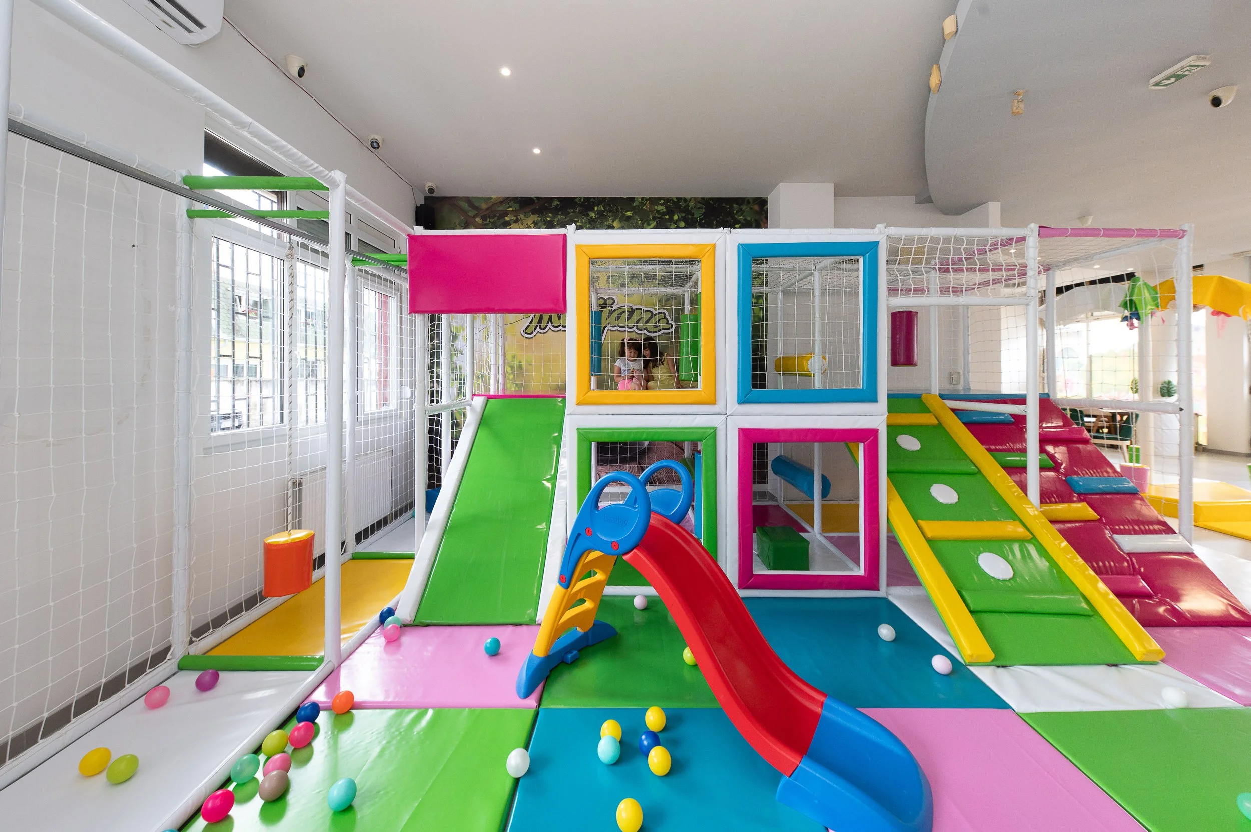 Colorful indoor children's play area with slides, climbing structures, and soft padding, enclosed by white safety netting.