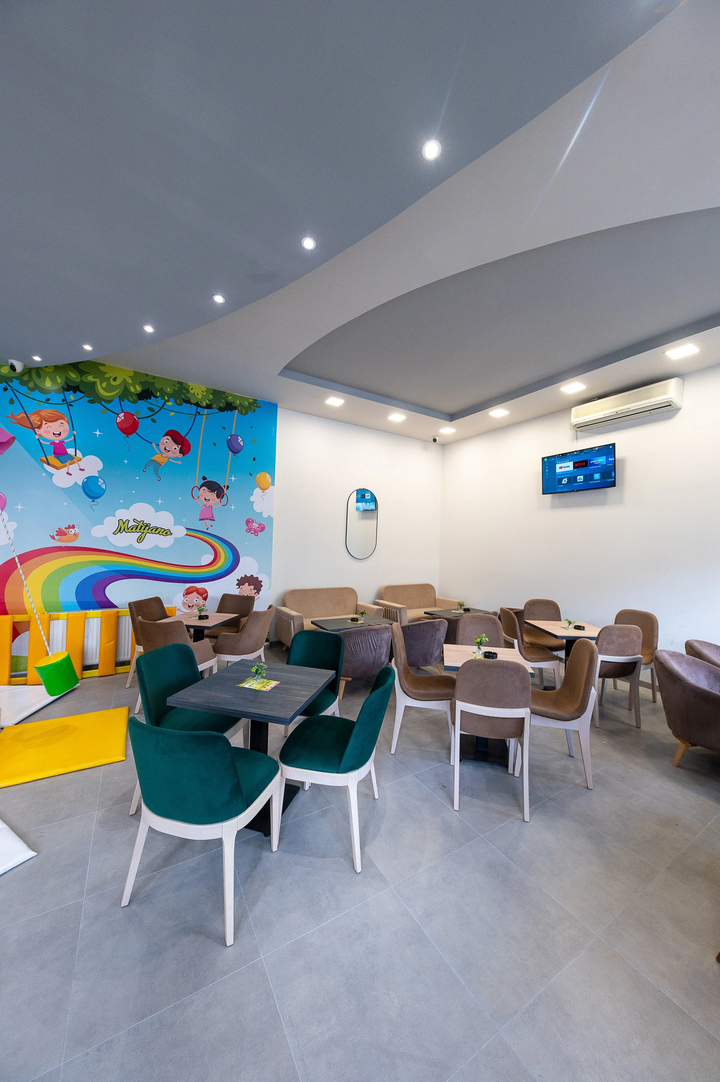 A cozy children’s play area or cafe with a colorful cartoon mural on the wall depicting kids on swings and rainbows, pastel-colored chairs, small tables, and a play mat with gym mats and padding for kids' safety.