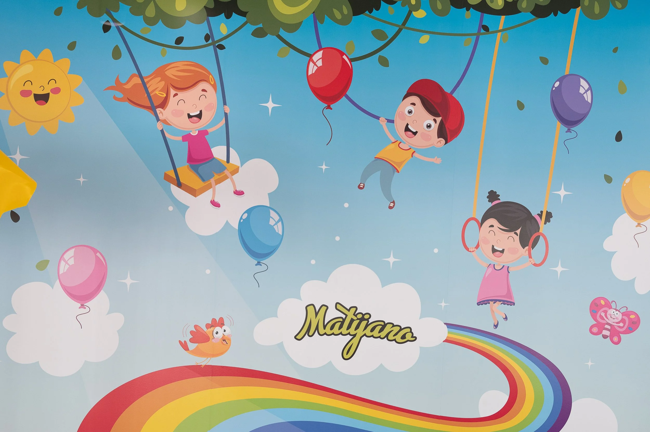 Colorful illustration of children playing on swings in the sky, surrounded by clouds, balloons, a smiling sun, a rainbow, and a fluffy pink butterfly. The name 'Matijano' is written on a cloud at the bottom.