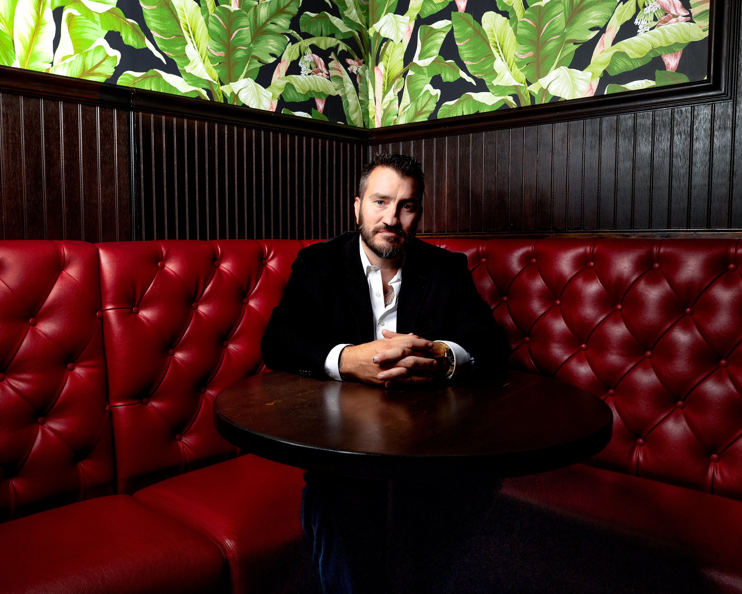 A man with a beard and mustache sitting at a dark wooden table in a booth with red leather upholstery. He is wearing a black blazer and a white shirt, and has his hands clasped on the table. Behind him is a wall with a botanical-themed mural of large green leaves.