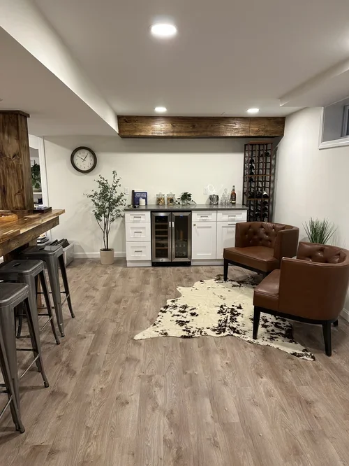 A cozy basement bar area with a small white cabinet, a potted plant, two brown armchairs, a cream and black patterned rug, and a wine rack, illuminated by ceiling lights.