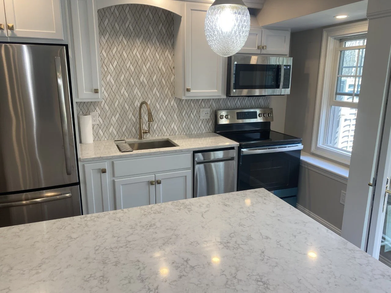Modern kitchen with white cabinets, marble countertops, stainless steel appliances, a black stove, microwave, and refrigerator, with a geometric backsplash and a large window.