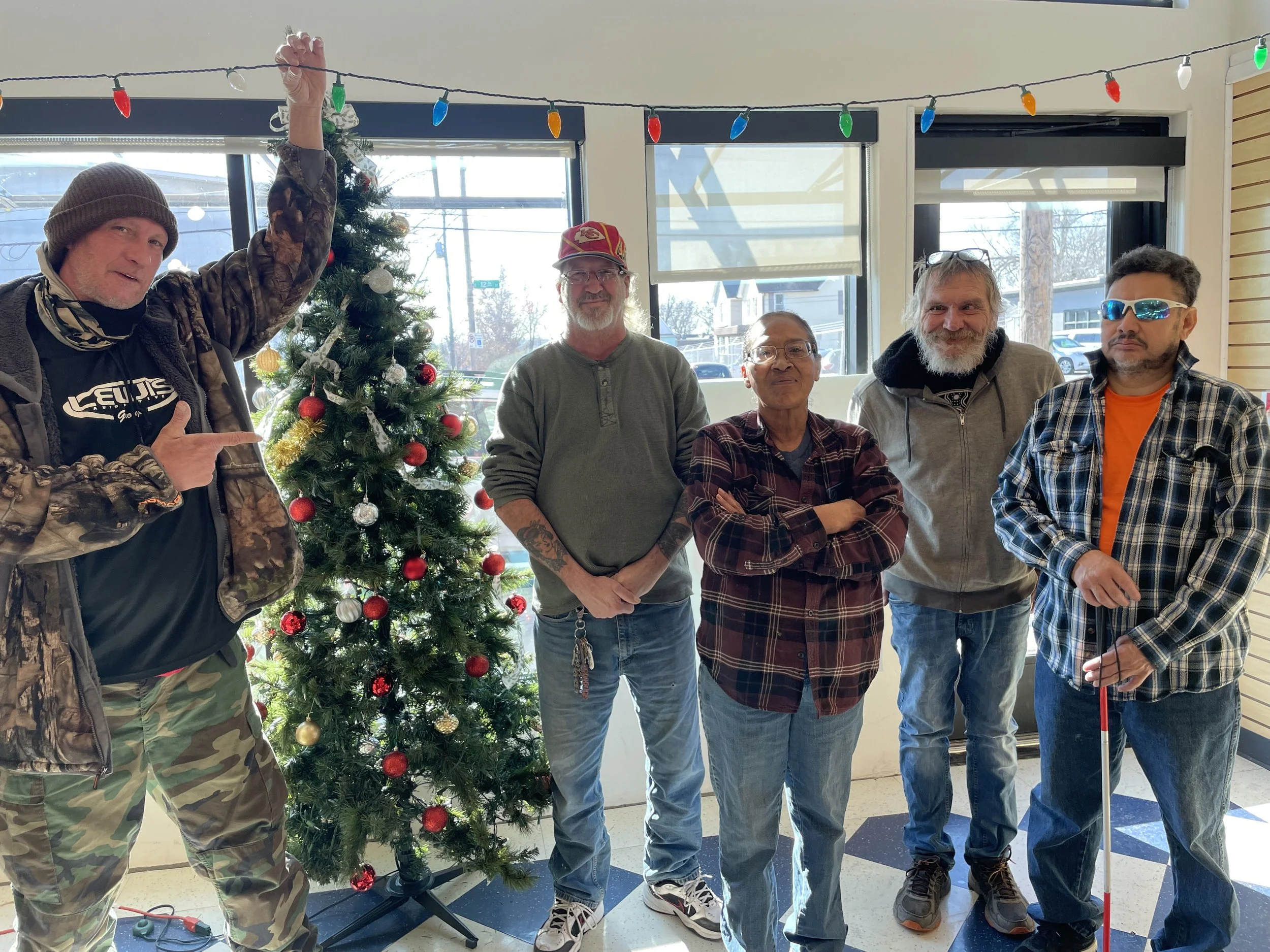 Group of six people standing inside near a decorated Christmas tree, with one person pointing at the tree, during the daytime.