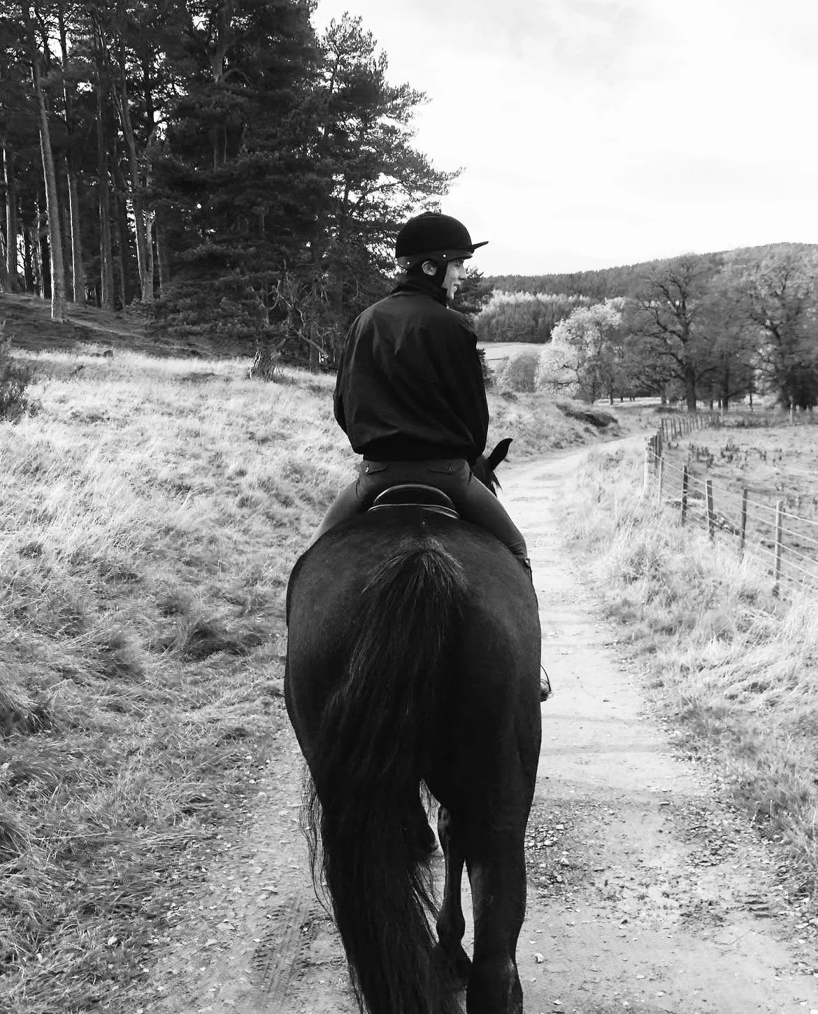 A person riding a horse on a dirt path through a rural landscape with trees and grassy fields in black and white.